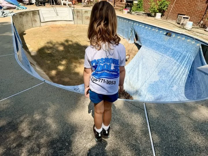A young girl standing in front of a broken swimming pool, with pool covers and debris inside. She is wearing a white T-shirt with blue lettering for a construction company, dark shorts, and black shoes with white socks.