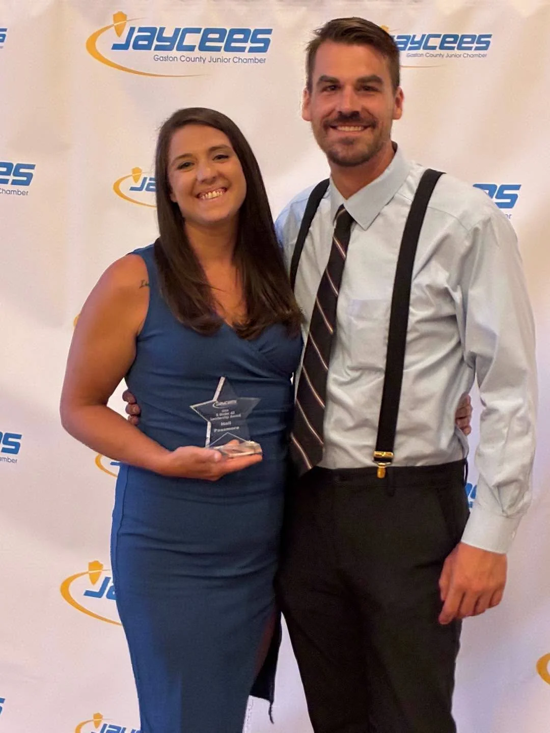 A woman in a blue dress holding a glass award and a man in a white shirt with suspenders standing together at an event with a 'JAYCEES' step and repeat backdrop.