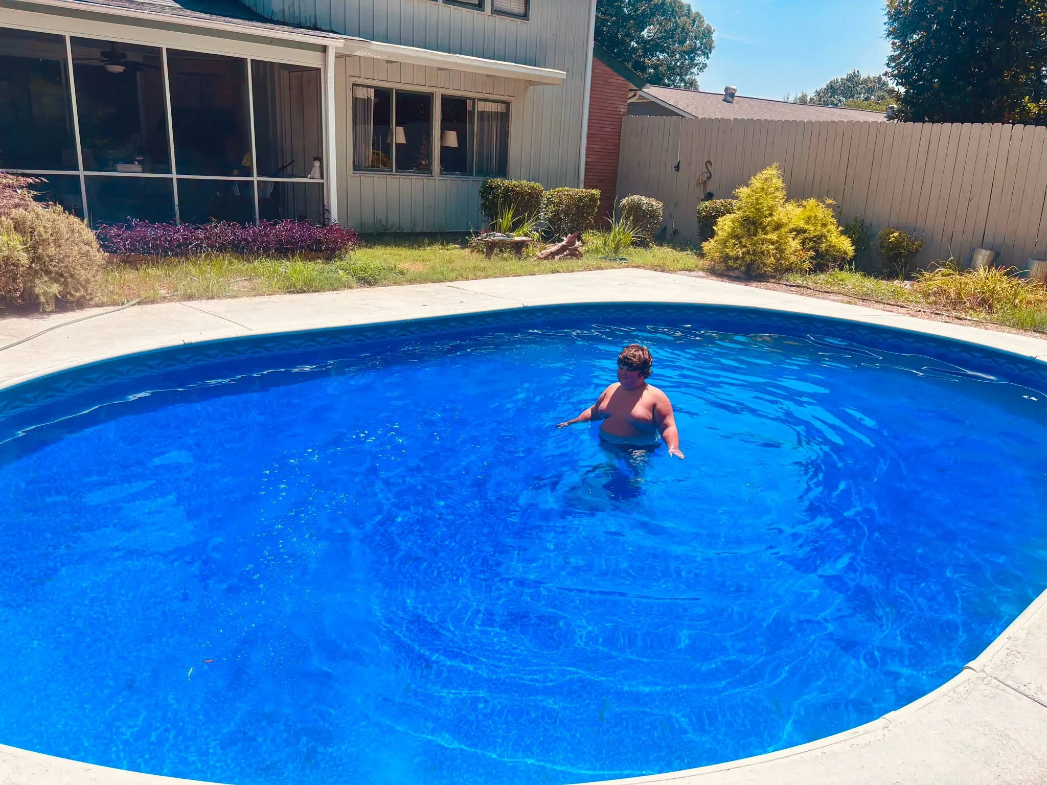 A backyard with a swimming pool, a person standing in the pool, and a fence with bushes and trees behind it.