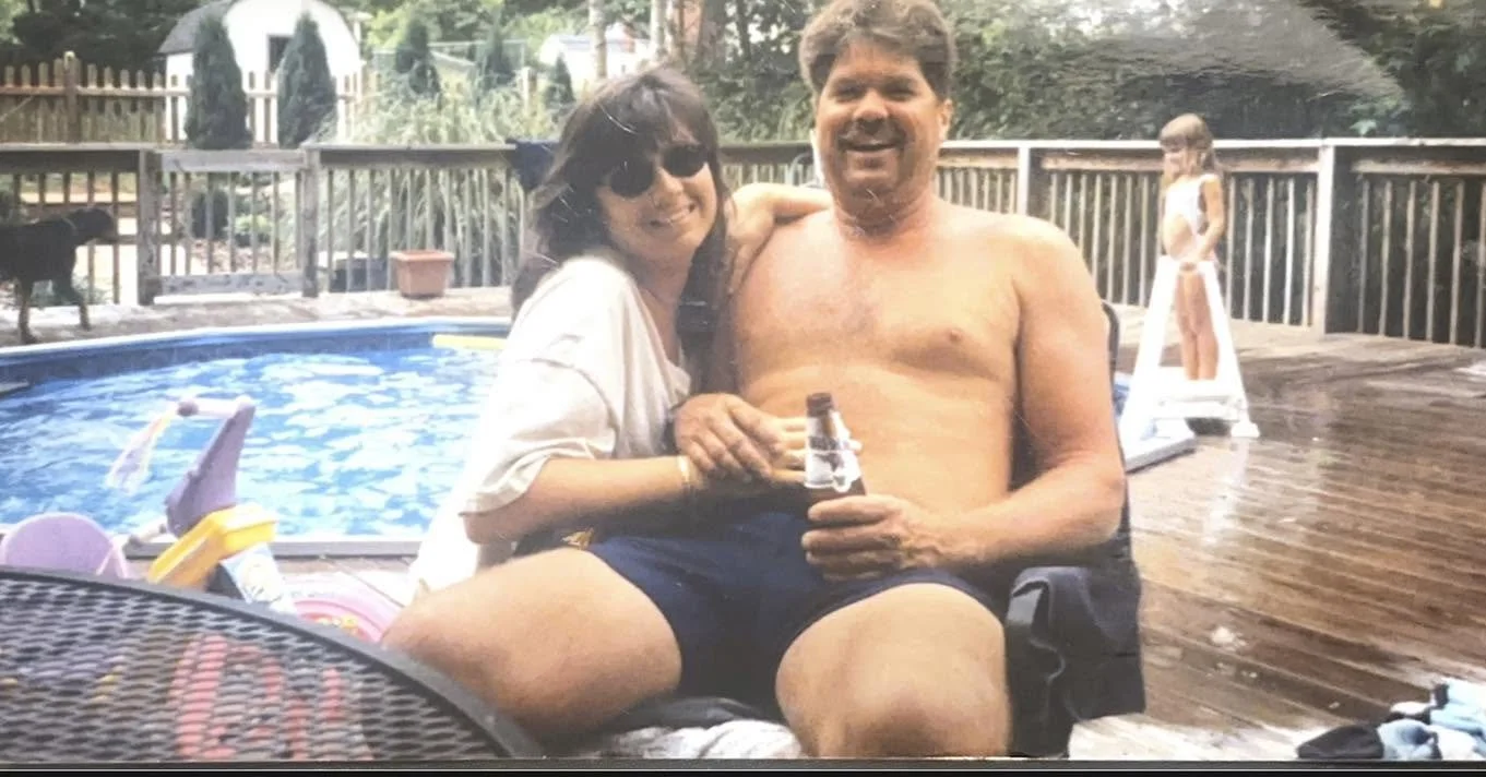A woman and a man sitting together on a deck near a swimming pool, holding a drink, with a young girl in the background on a slide.