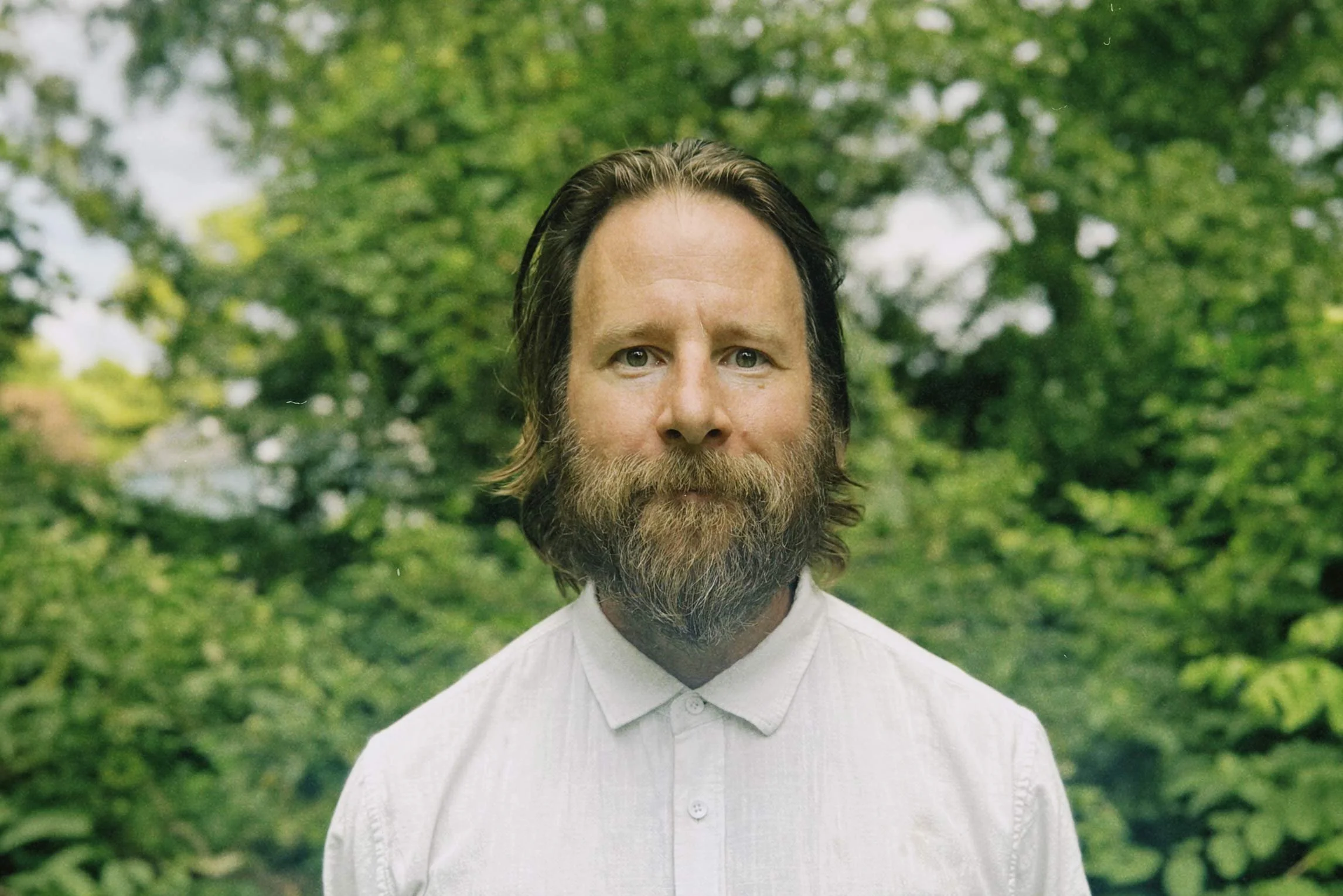 A middle-aged man with a beard and long brown hair, wearing a white button-up shirt, standing outdoors in front of green trees with a cloudy sky in the background.