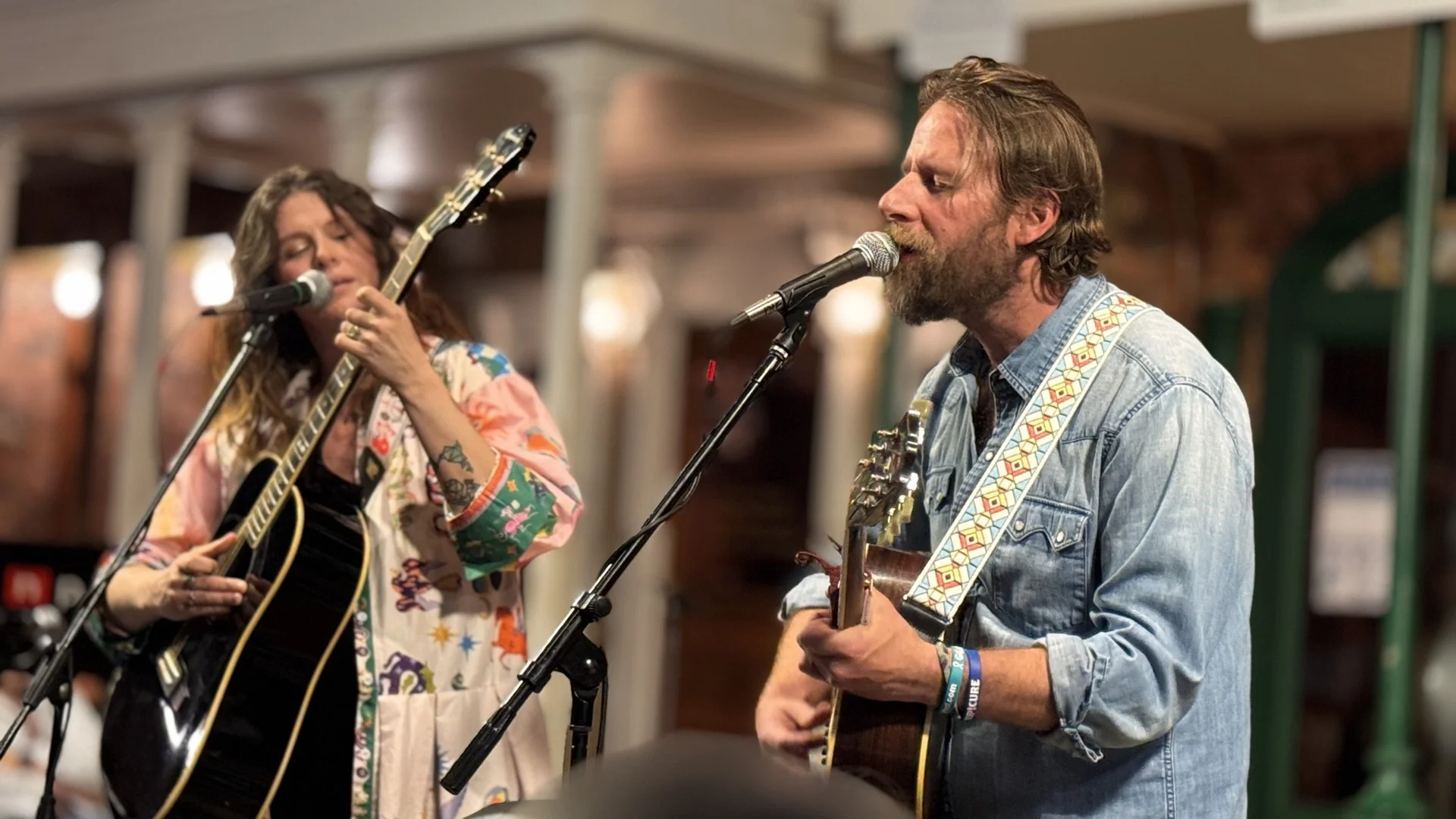 A woman and a man are performing together with guitars in a cozy indoor setting. The woman has long, curly hair and is wearing a colorful, patterned garment. The man has a beard and is wearing a denim shirt. Both are singing into microphones.