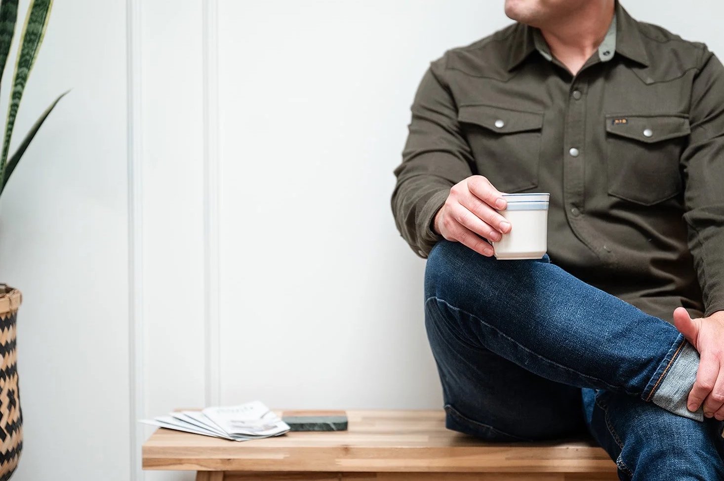 A man sitting on a wooden bench, wearing a green shirt and jeans, holding a white mug with a blue stripe, with papers on the bench and a potted plant nearby.