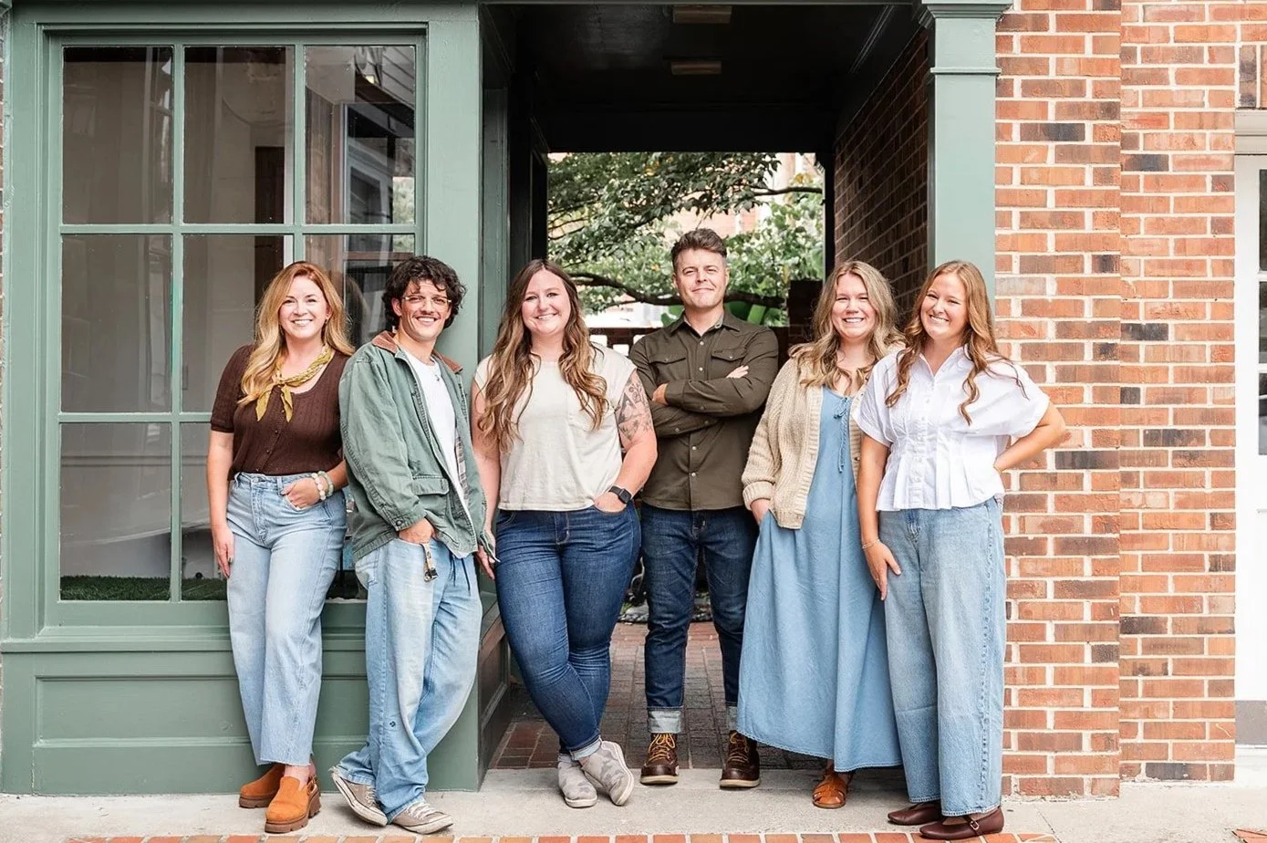 Group of six young adults standing outside a brick and green storefront, smiling at the camera.