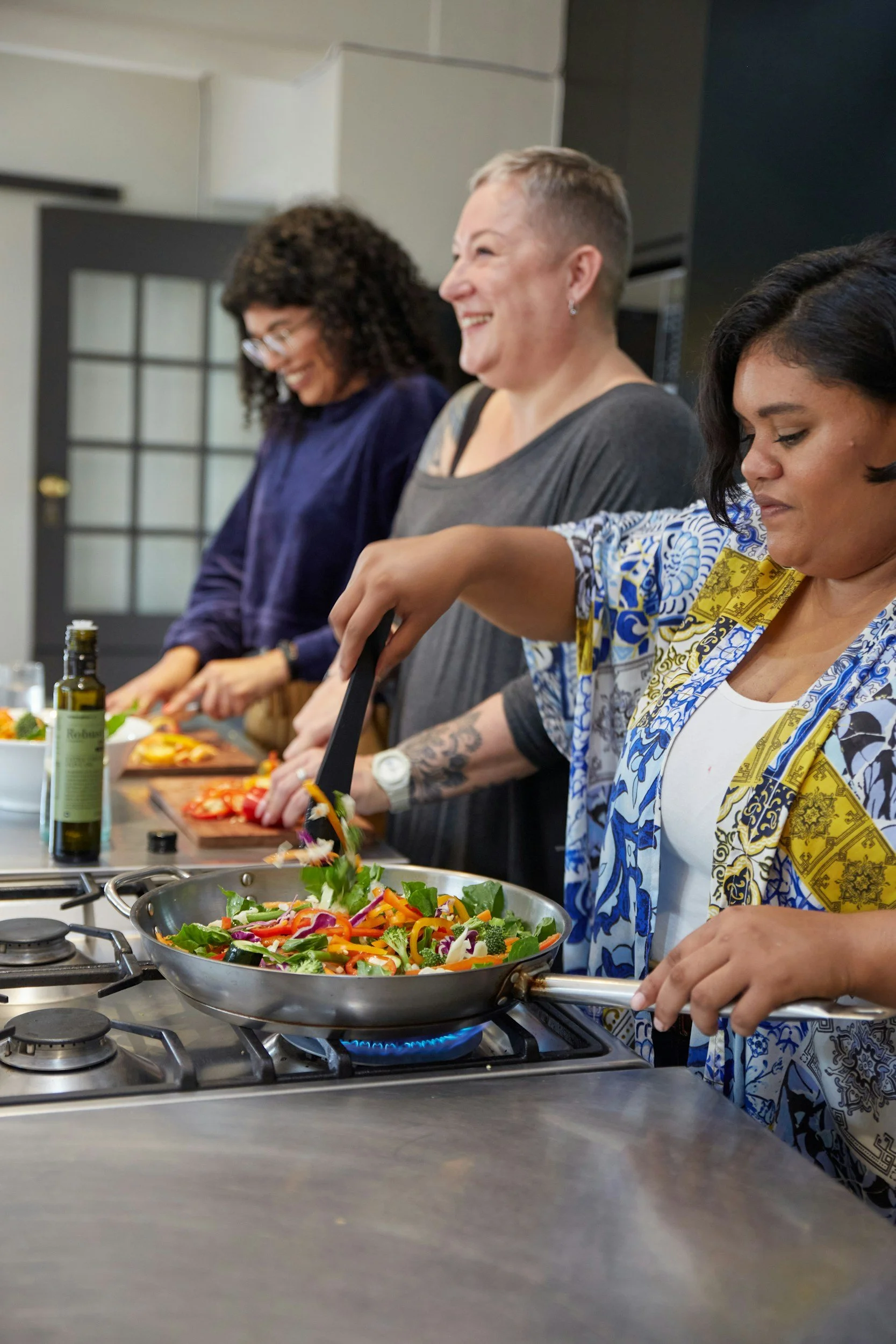 Three women cooking and preparing a vegetable salad in a kitchen. One woman is stirring vegetables in a pan on the stove, and two others are chopping vegetables at the counter.