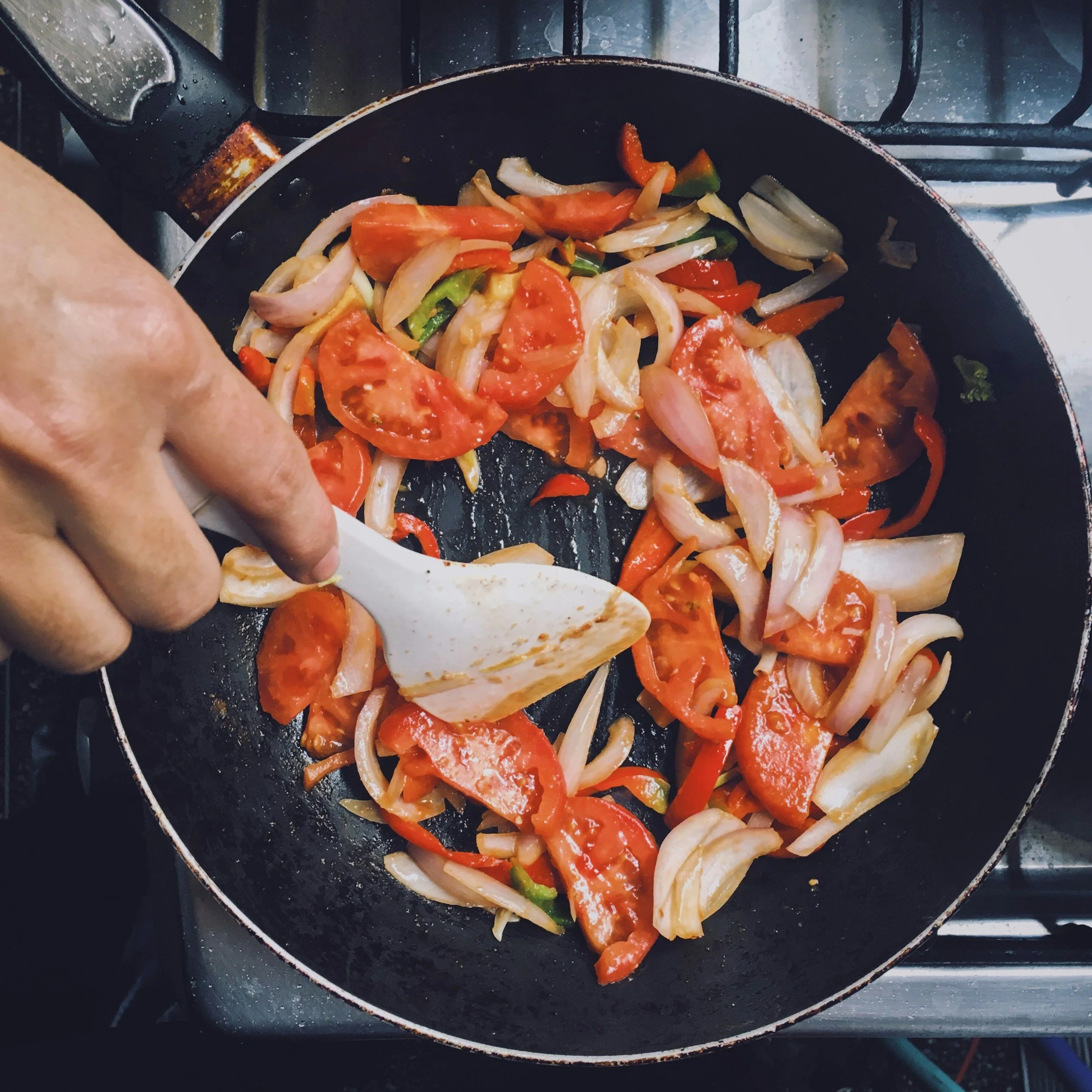 Sautéing chopped onions, tomatoes, and bell peppers in a black non-stick skillet on a stove with a silver handle.