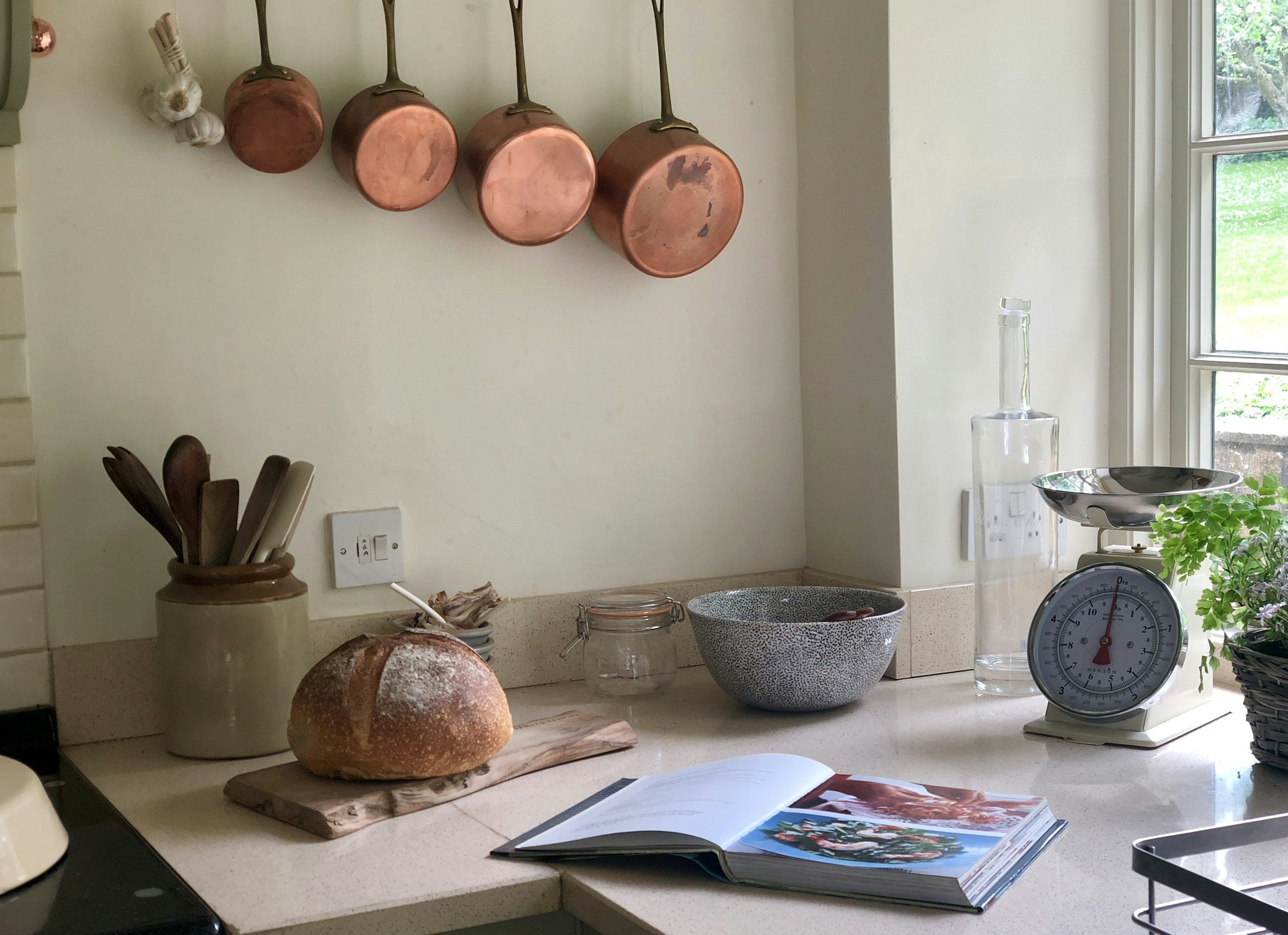 A kitchen countertop with a loaf of bread on a wooden cutting board, a bowl, a jar, a clock, a tall glass bottle, and a potted plant near a window, with copper pots hanging on the wall.