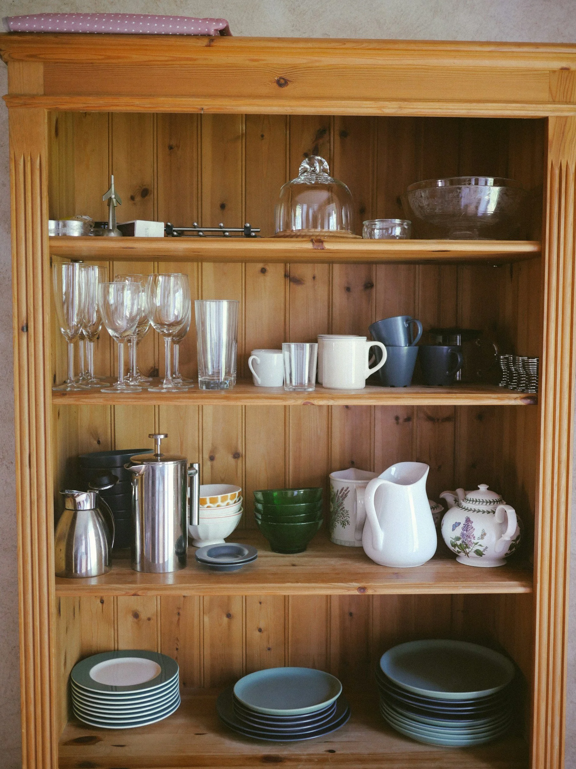 Empty wooden cabinet with glassware, dishes, mugs, bowls, and kitchen utensils arranged on the shelves.