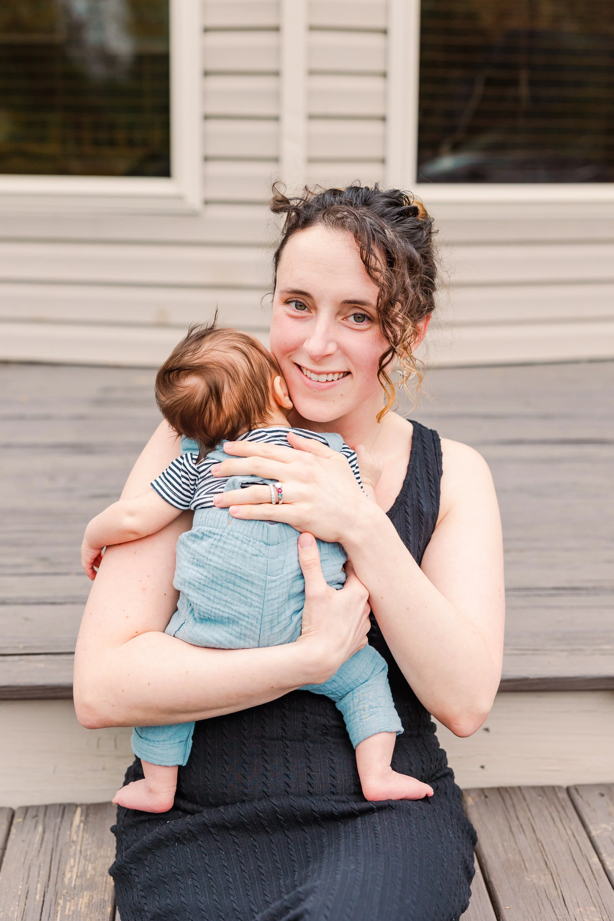 A smiling woman with curly dark hair holding a small child with light brown hair in front of a house with beige siding and a wooden deck.