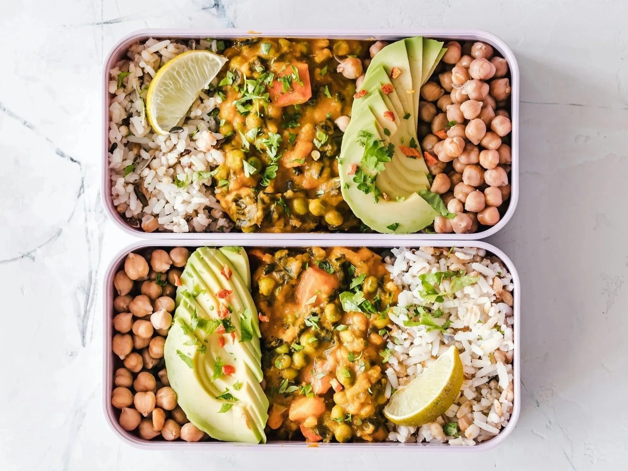 Top view of a lunch container holding cooked rice, chickpeas, sliced avocado, and a vegetable curry garnished with chopped cilantro and red pepper flakes, with lemon wedges on the side.