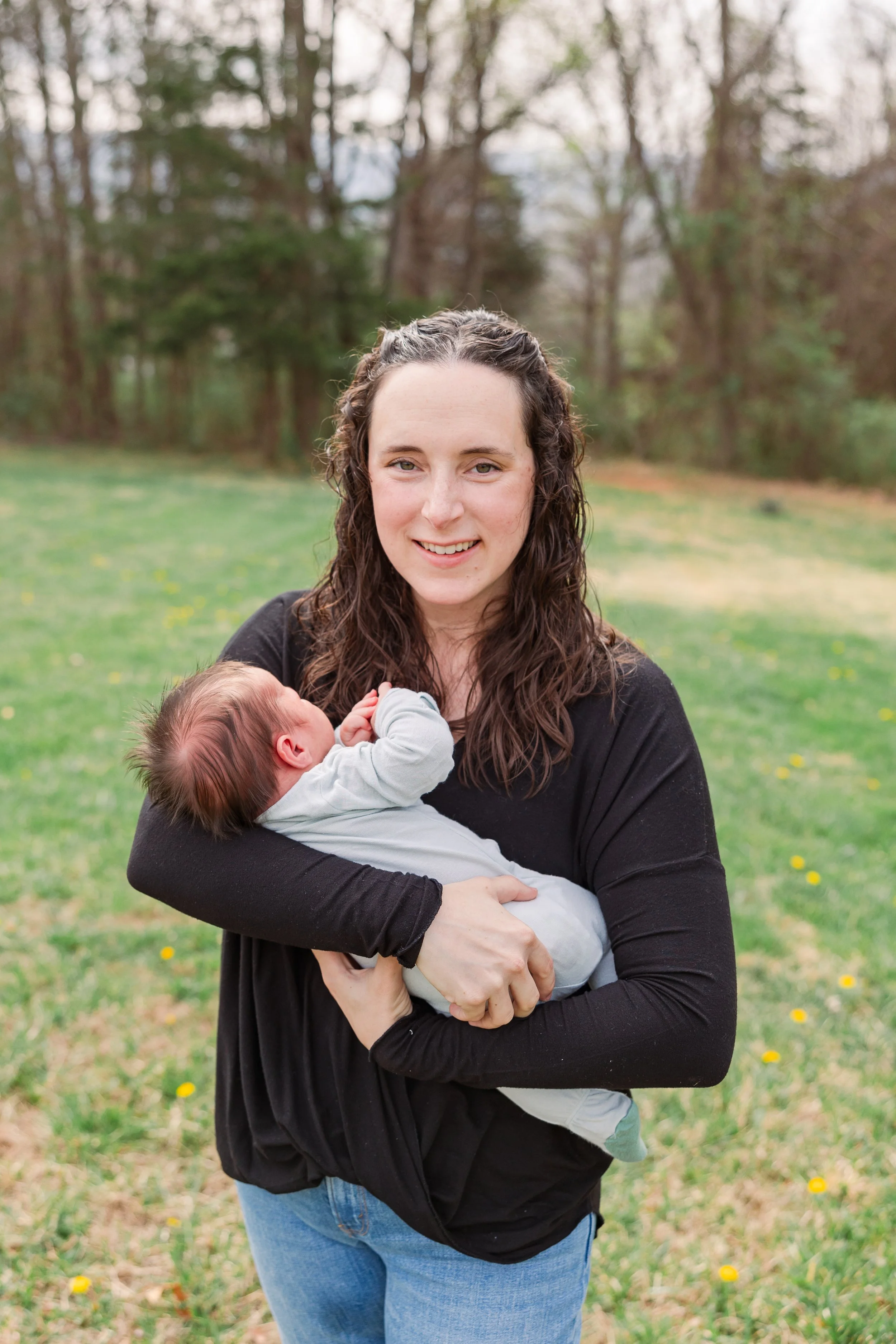 A woman with long curly hair smiling while holding a baby in an outdoor park with trees and grass.