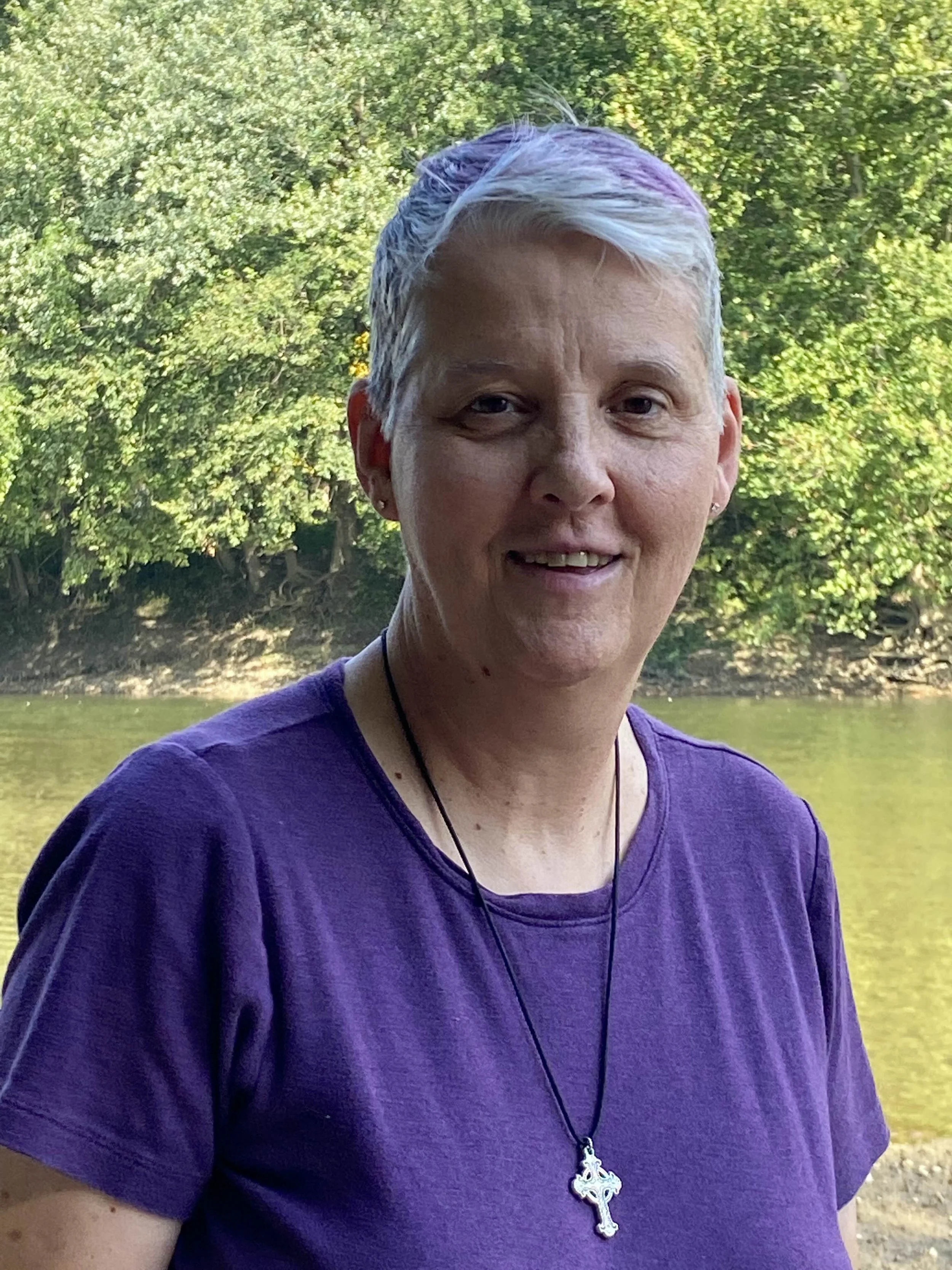 A woman with short white hair wearing a purple shirt, a necklace with a cross pendant, and ear earrings, standing outdoors near a body of water with green trees in the background.