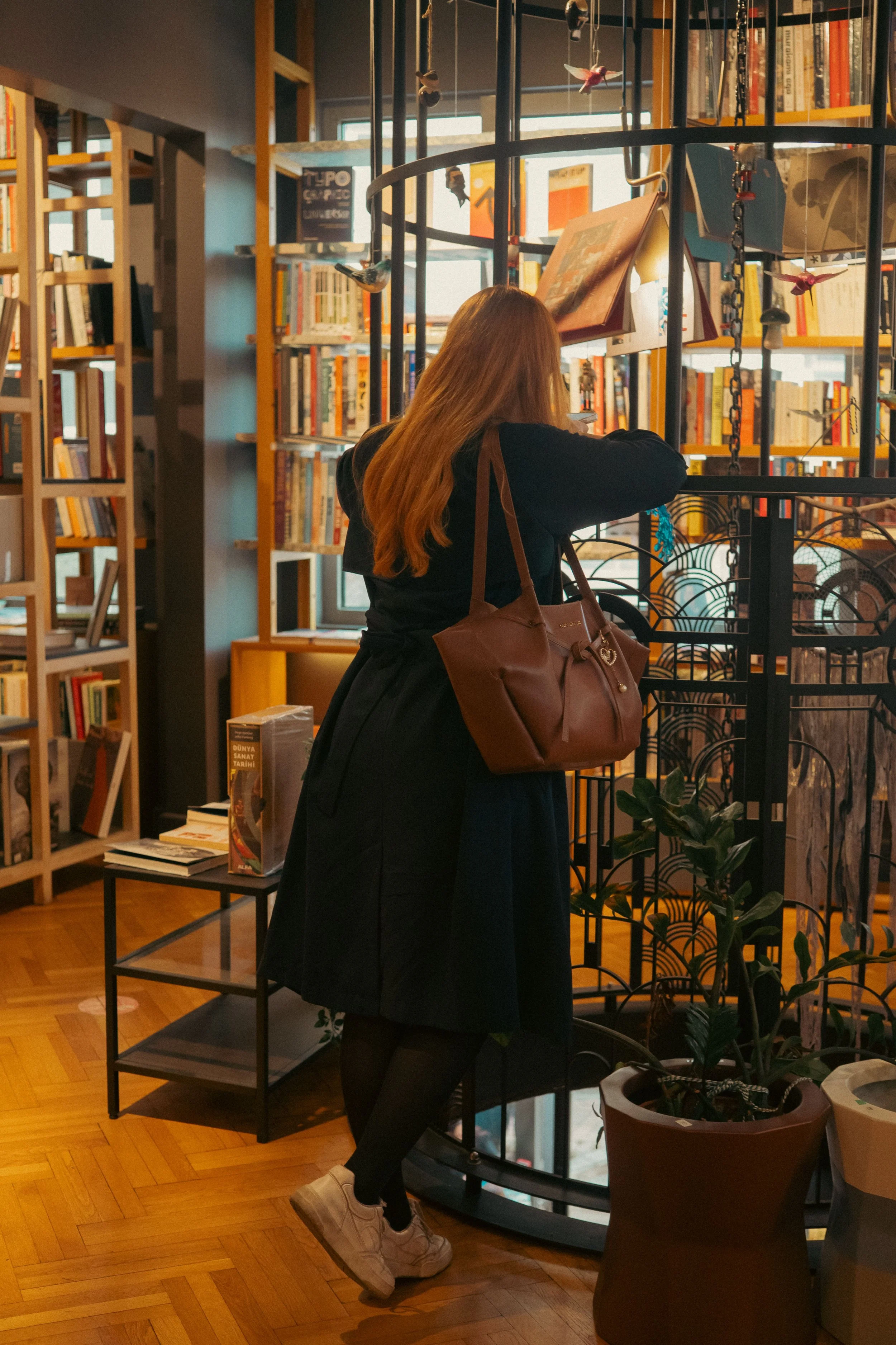 A woman with long hair wearing a black coat, black pants, and white sneakers is standing in a bookstore, looking at the shelves. She has a large brown purse on her shoulder. The bookstore has wooden floors, bookshelves filled with books, and decorative hanging ornaments.
