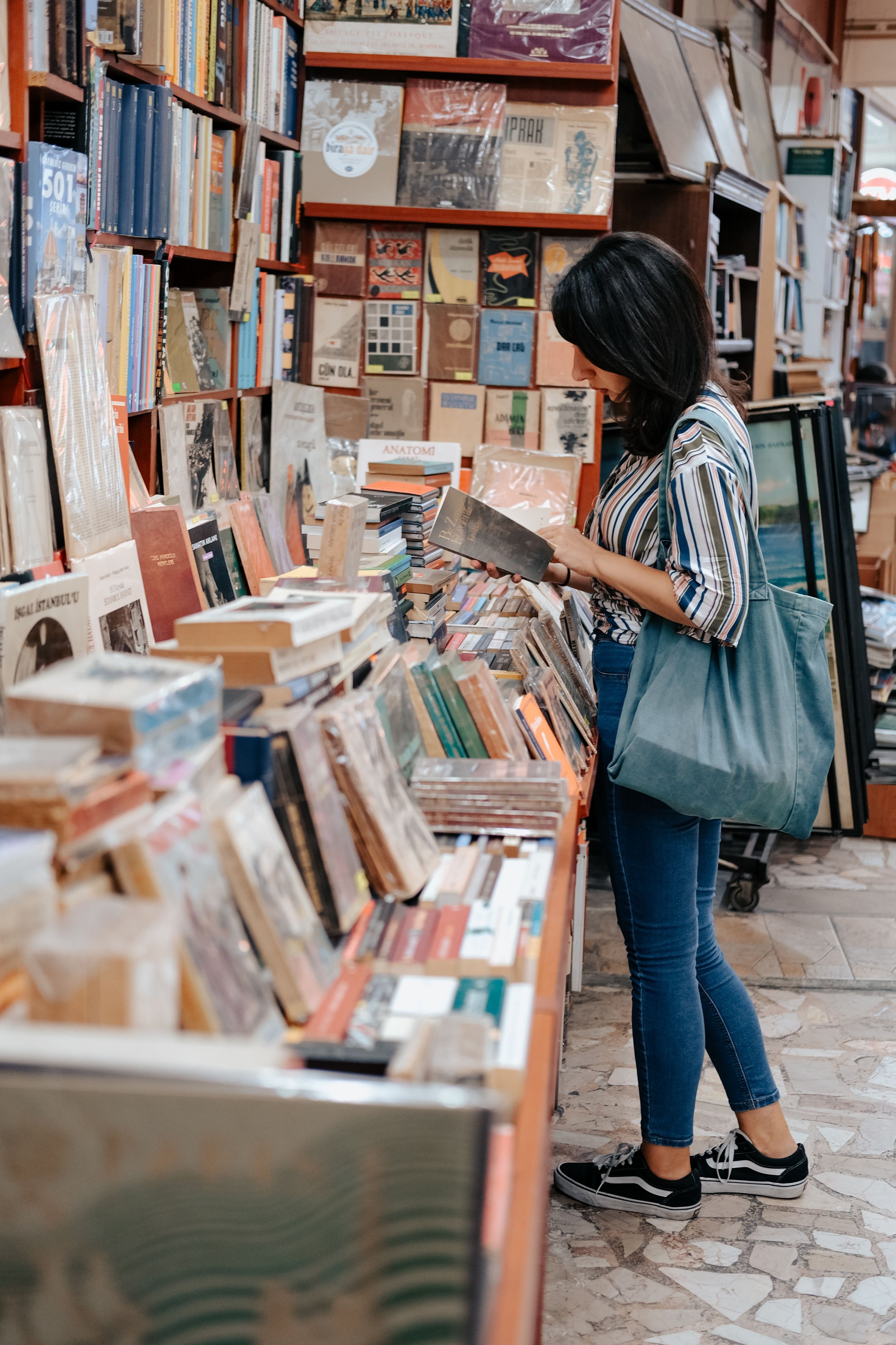 A woman with black hair, wearing a striped shirt and blue jeans, is browsing books in a bookstore or library. She has a large blue tote bag over her shoulder and is looking at a book in her hands. The background shows shelves filled with various books and artwork.
