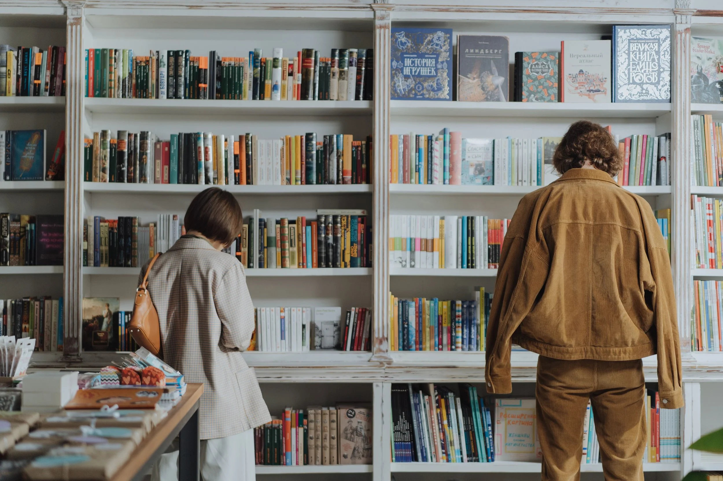 Two women browsing bookshelves in a bookstore or library, one with short hair and a checkered coat, the other with curly hair and a brown jacket.