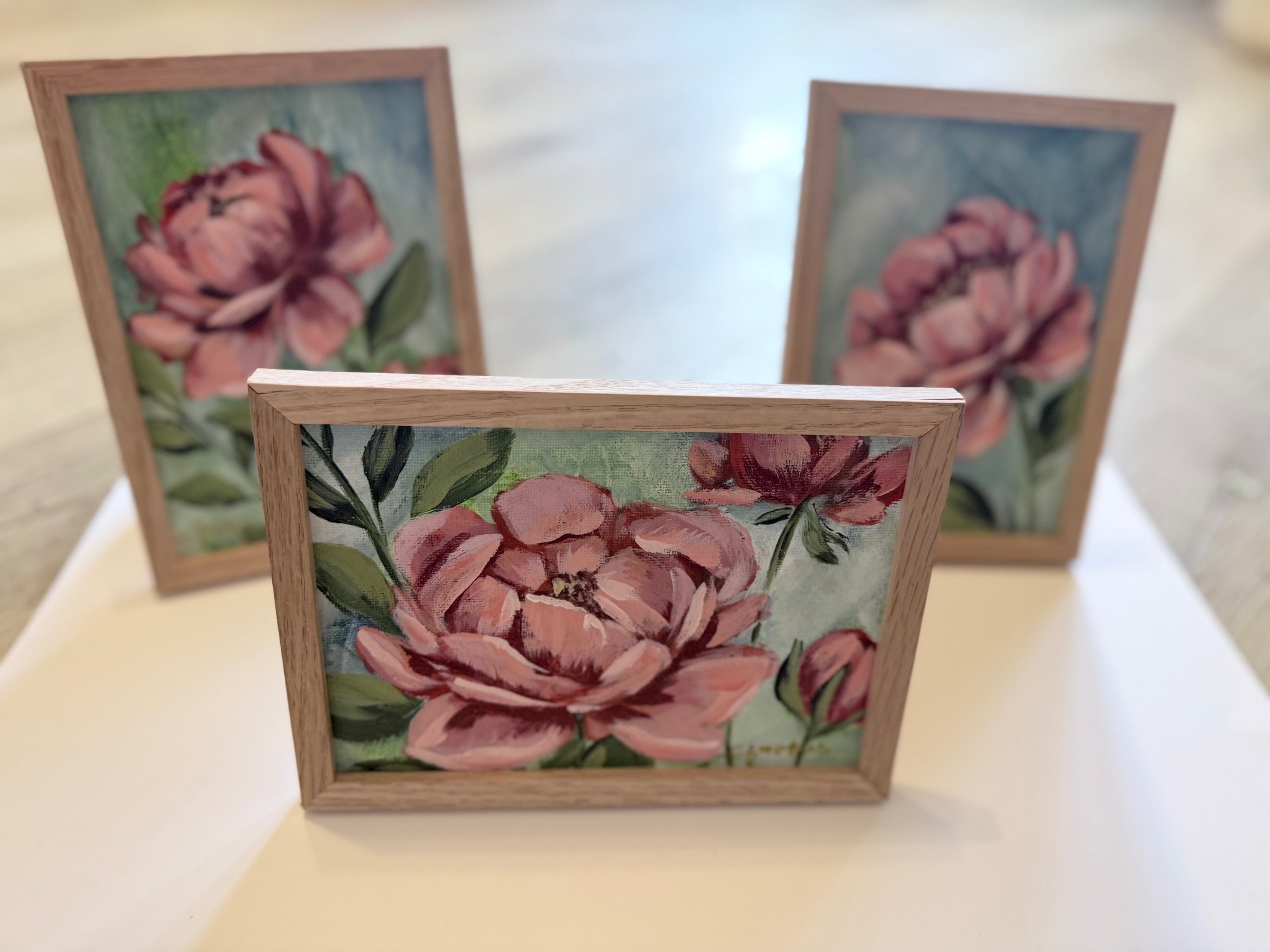 Three framed paintings of pink roses with green leaves, displayed on a table or shelf.