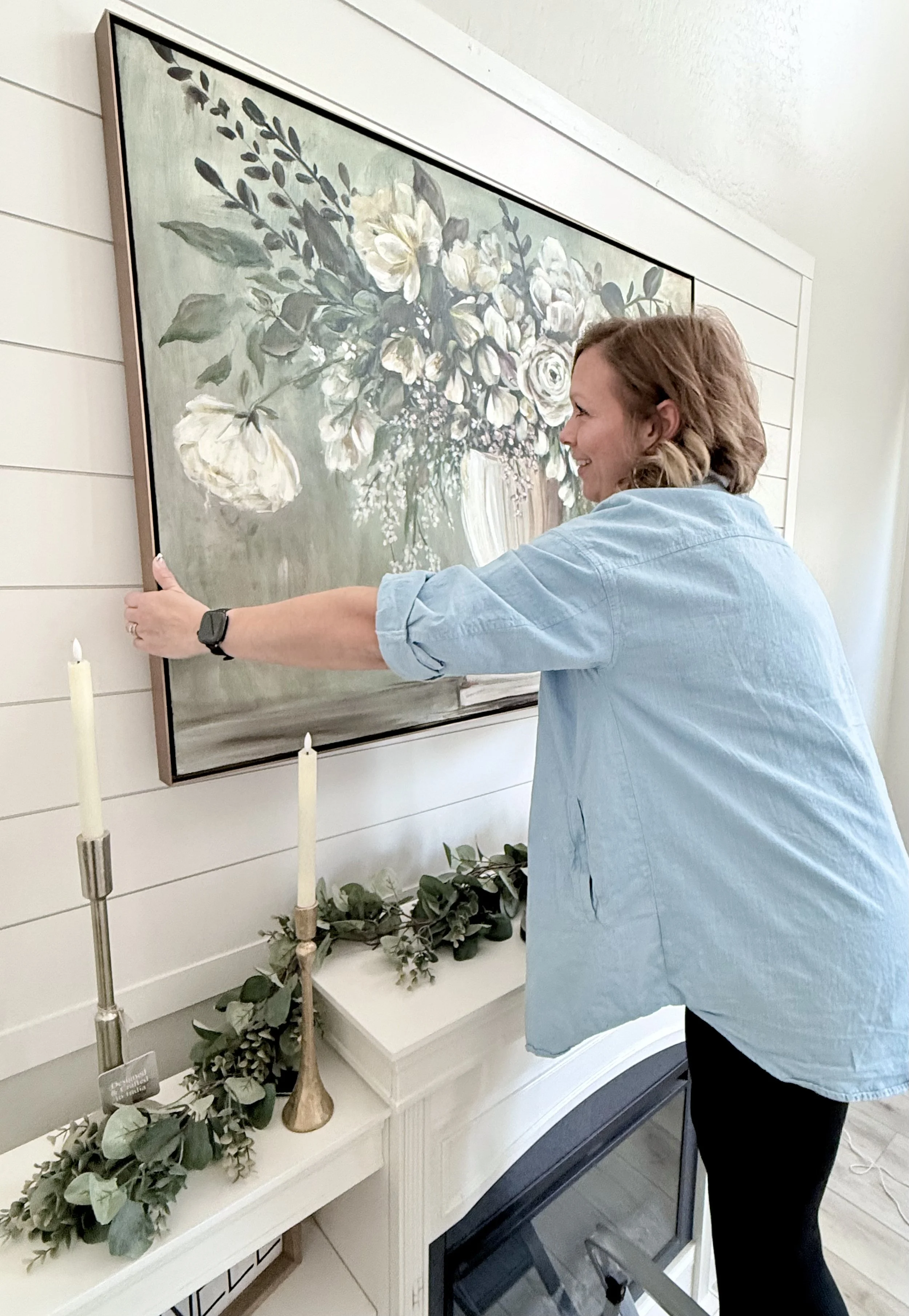 A woman wearing a light blue shirt and black pants placing a frame on a wall above a fireplace mantel. The mantel is decorated with artificial green leafy garland, tall candles, and a small sign. A large floral painting with white and beige flowers is hung on the wall.