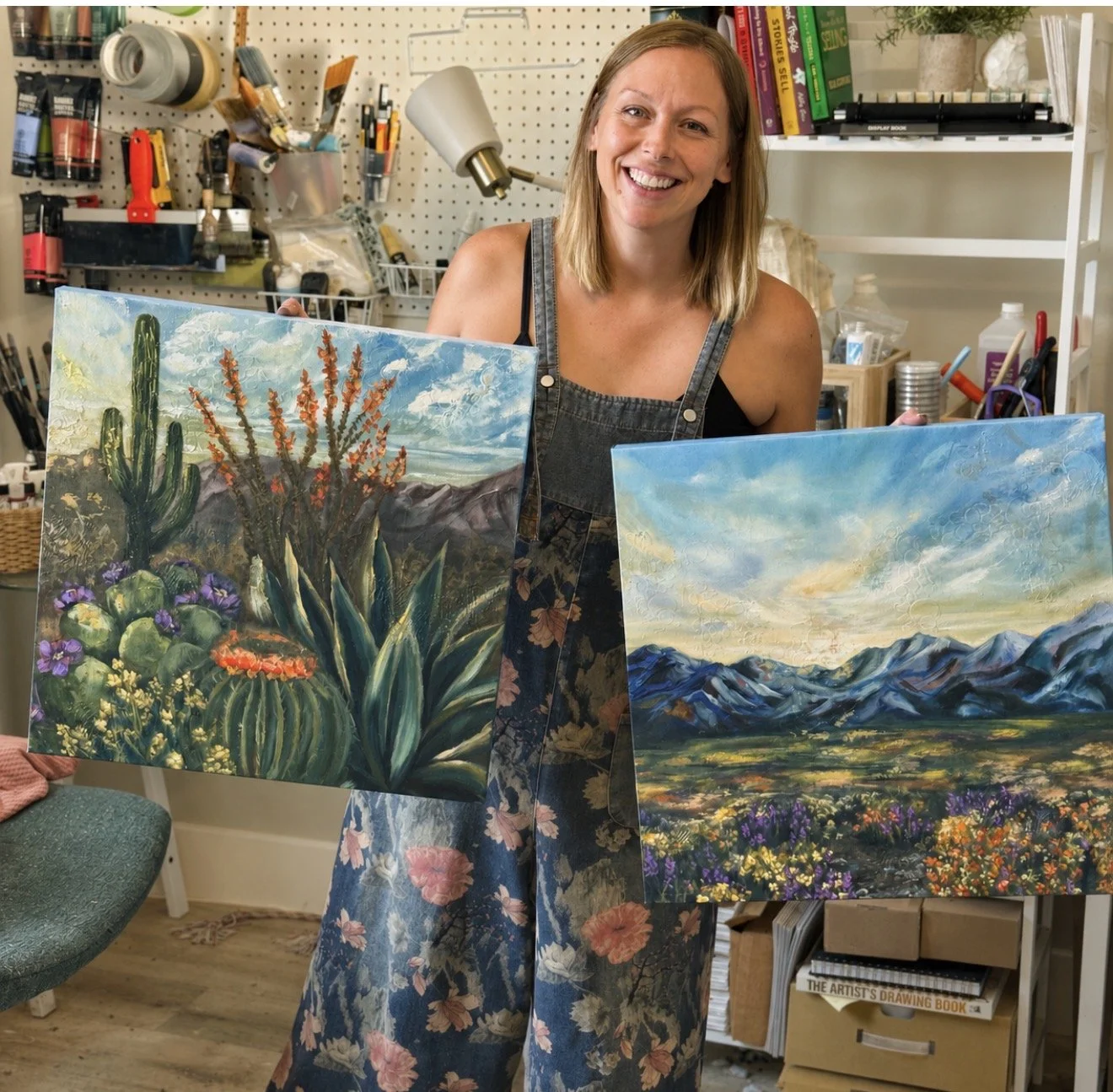 A woman smiling and holding two landscape paintings, one depicting a desert scene with cacti and succulents, and the other showing mountains and flowers, inside an art studio with art supplies and books in the background.