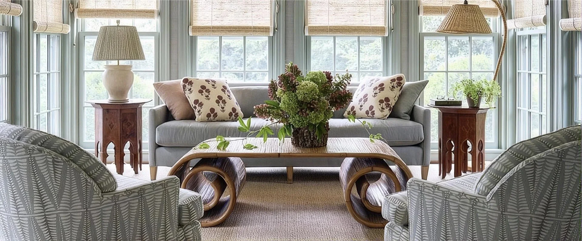 Living room with two patterned armchairs, a sofa with floral pillows, and a wooden coffee table with a large flower arrangement, illuminated by large windows and lamps on side tables.