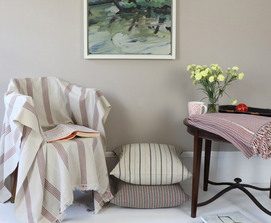 A cozy sitting area with a chair draped in a beige striped blanket, an open book on it, two striped floor cushions, a small wooden table with a red and white patterned cloth, a vase of yellow flowers, a mug, and a framed abstract painting on the wall.