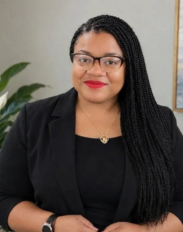 A woman with braided hair, wearing glasses, a black blazer, and a necklace, posing for a professional portrait indoors. counselor, parental burnout, childhood wounds, therapist for parents, parent alienation, PTSD wounds, trauma