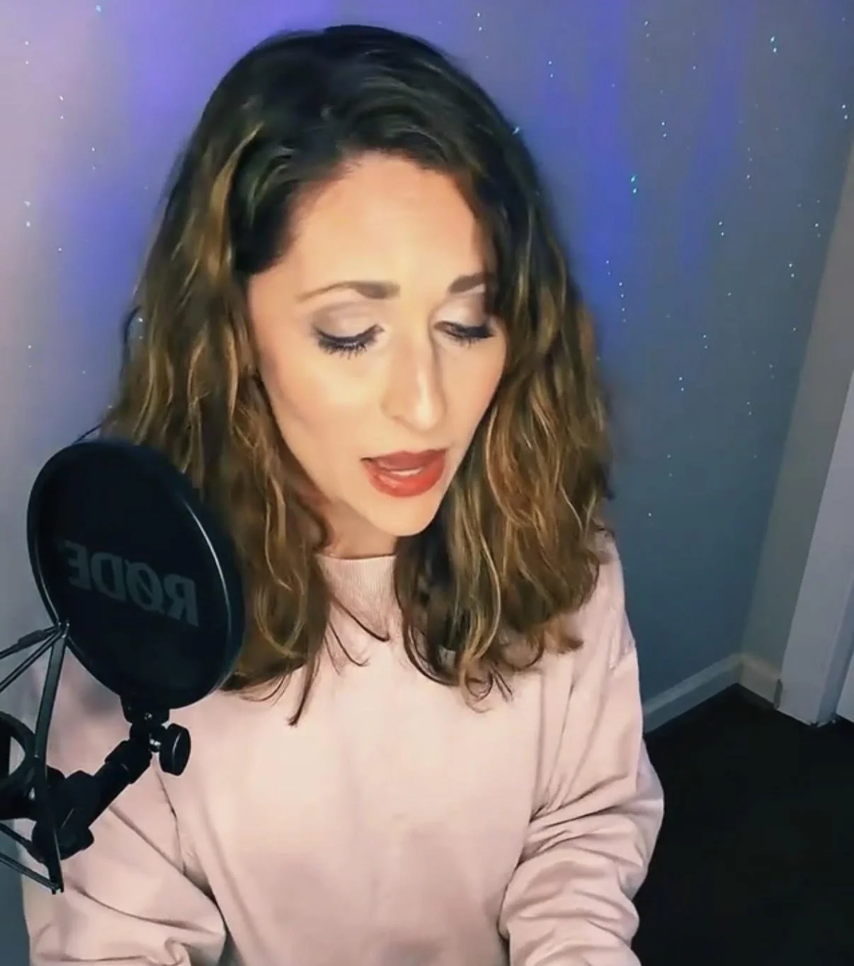 A woman singing into a Rode microphone in a home recording space, with a starry space-themed wall behind her.