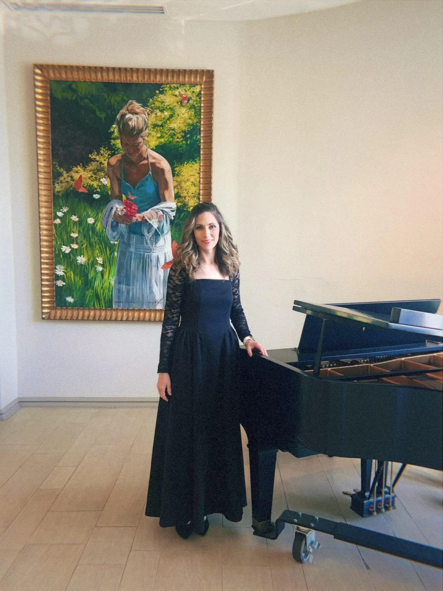 A woman in a black dress standing next to a black grand piano in a room with beige walls and wooden flooring. There is a colorful painting of a woman with short hair in a blue top holding a bouquet of flowers hung on the wall behind her.