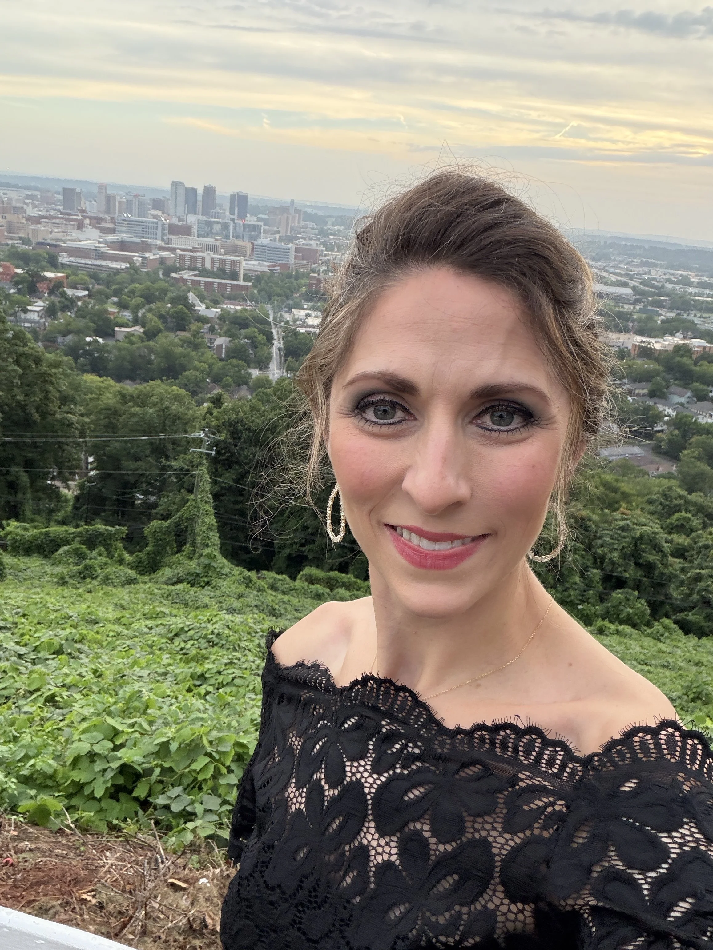 Close-up of a smiling woman with blue eyes and light makeup, wearing earrings and a black lace off-the-shoulder dress, standing outdoors with a cityscape in the background, including green trees and buildings, during mixed cloudy weather.