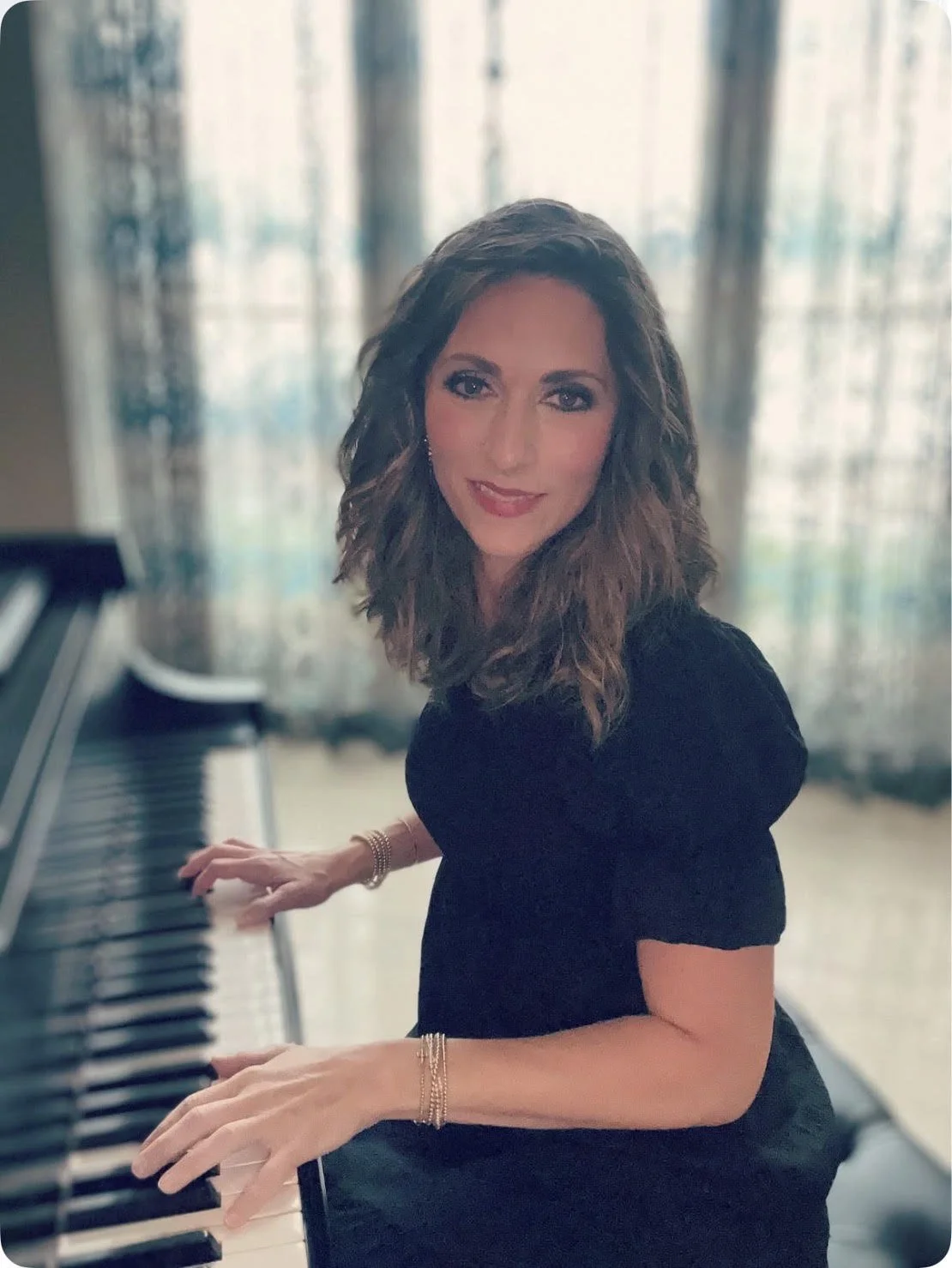 A woman with wavy brown hair, wearing a black top, playing a grand piano indoors with sheer curtains and large windows behind her.
