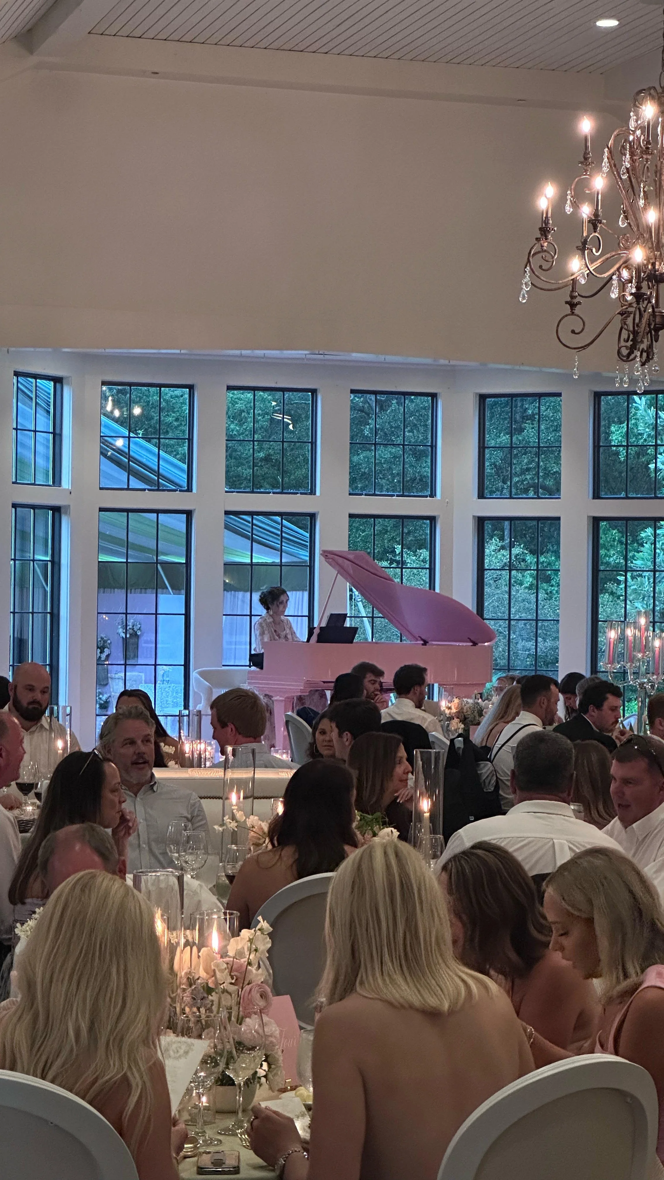 Elegant indoor event with guests seated at tables, floral centerpieces, and a musician playing a pink grand piano in front of large windows with greenery outside, chandeliers overhead.