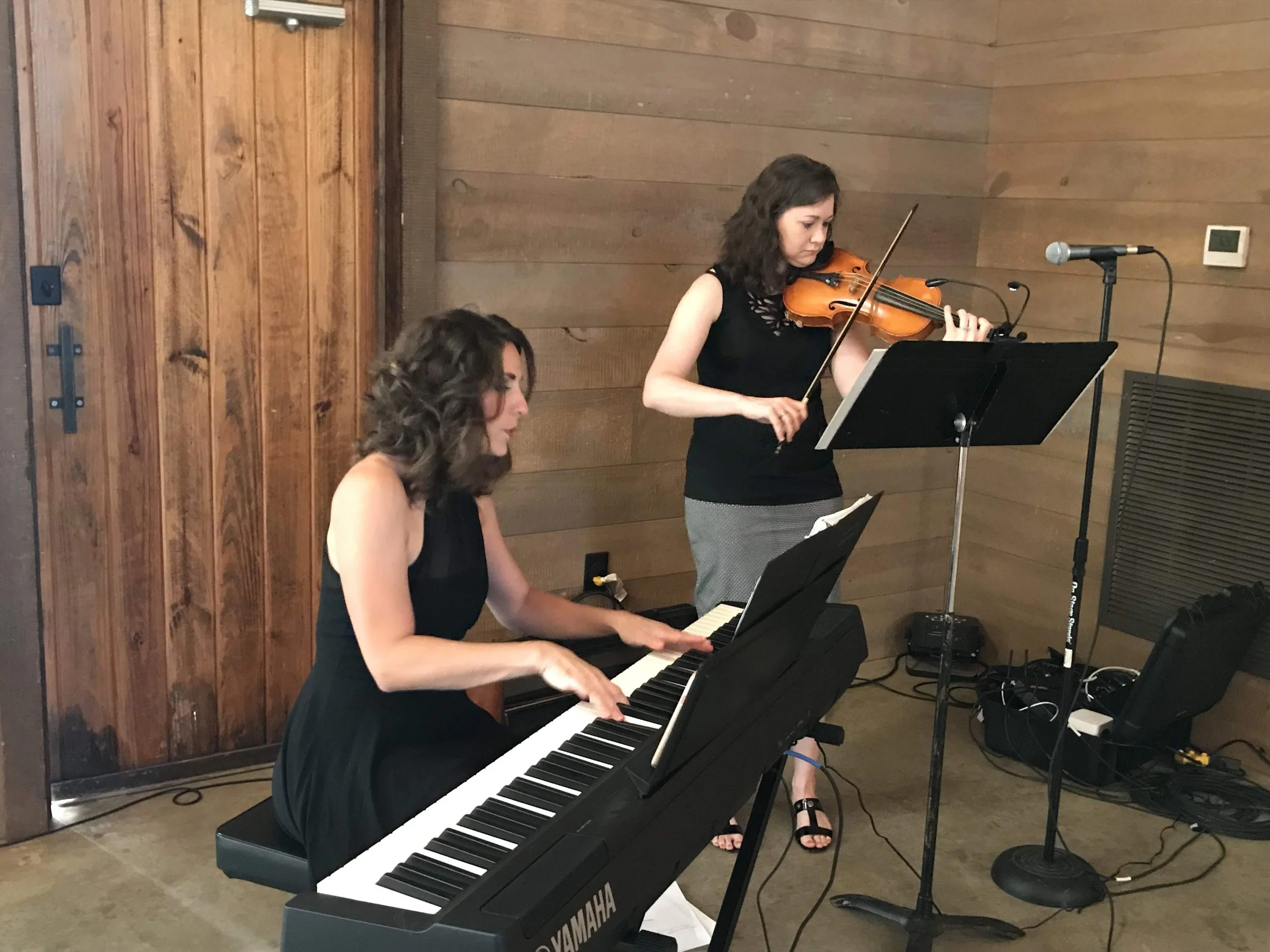Two women are performing music in a wooden room; one is playing a violin while the other is playing a keyboard piano.