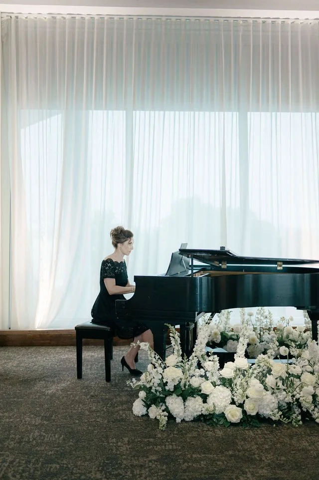 A woman in a black dress playing a grand piano in a bright room with large, white curtains and a floral arrangement in front of the piano.