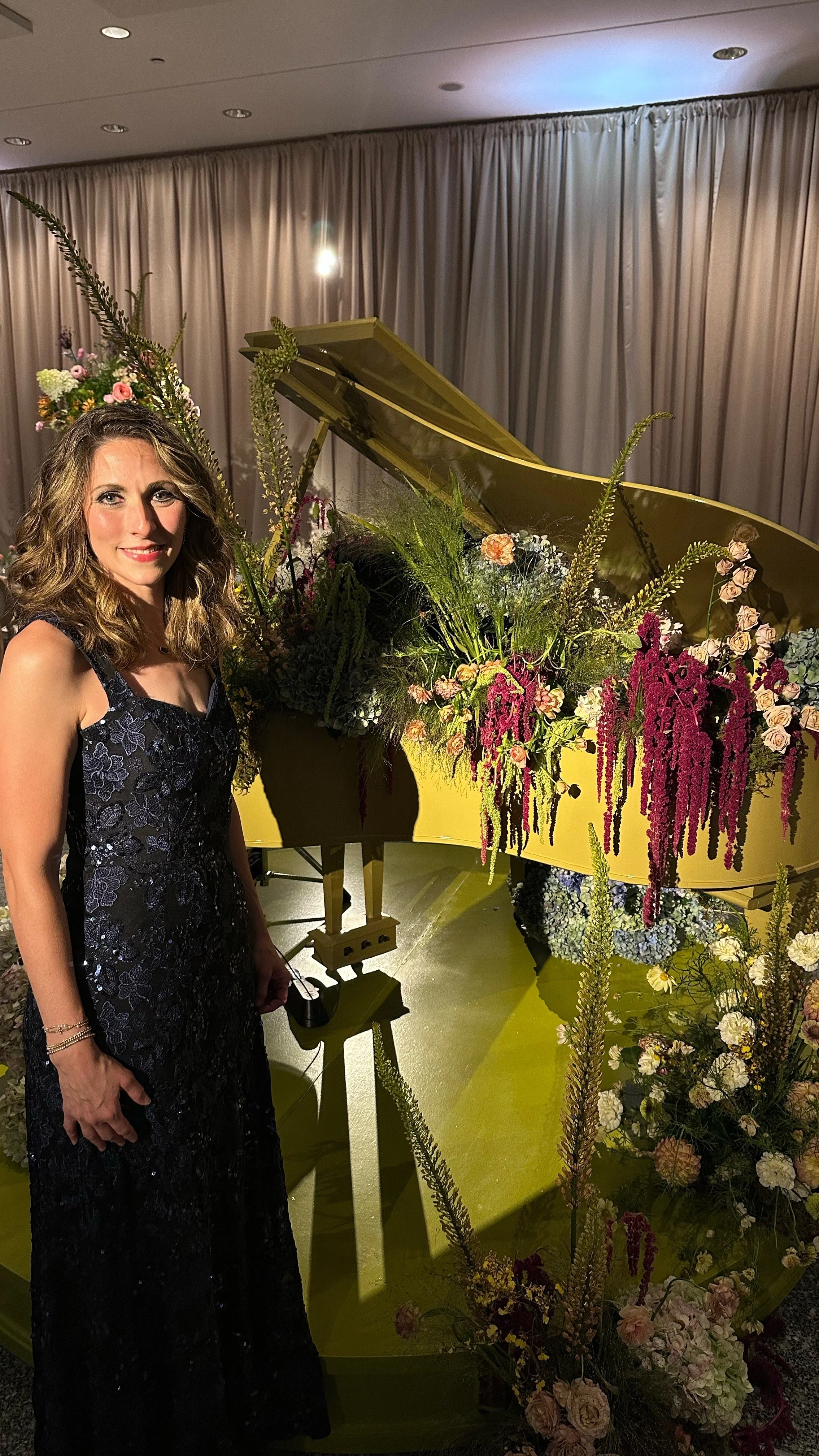 A woman in a black dress standing next to a yellow grand piano decorated with colorful flowers and greenery, with beige curtains in the background.