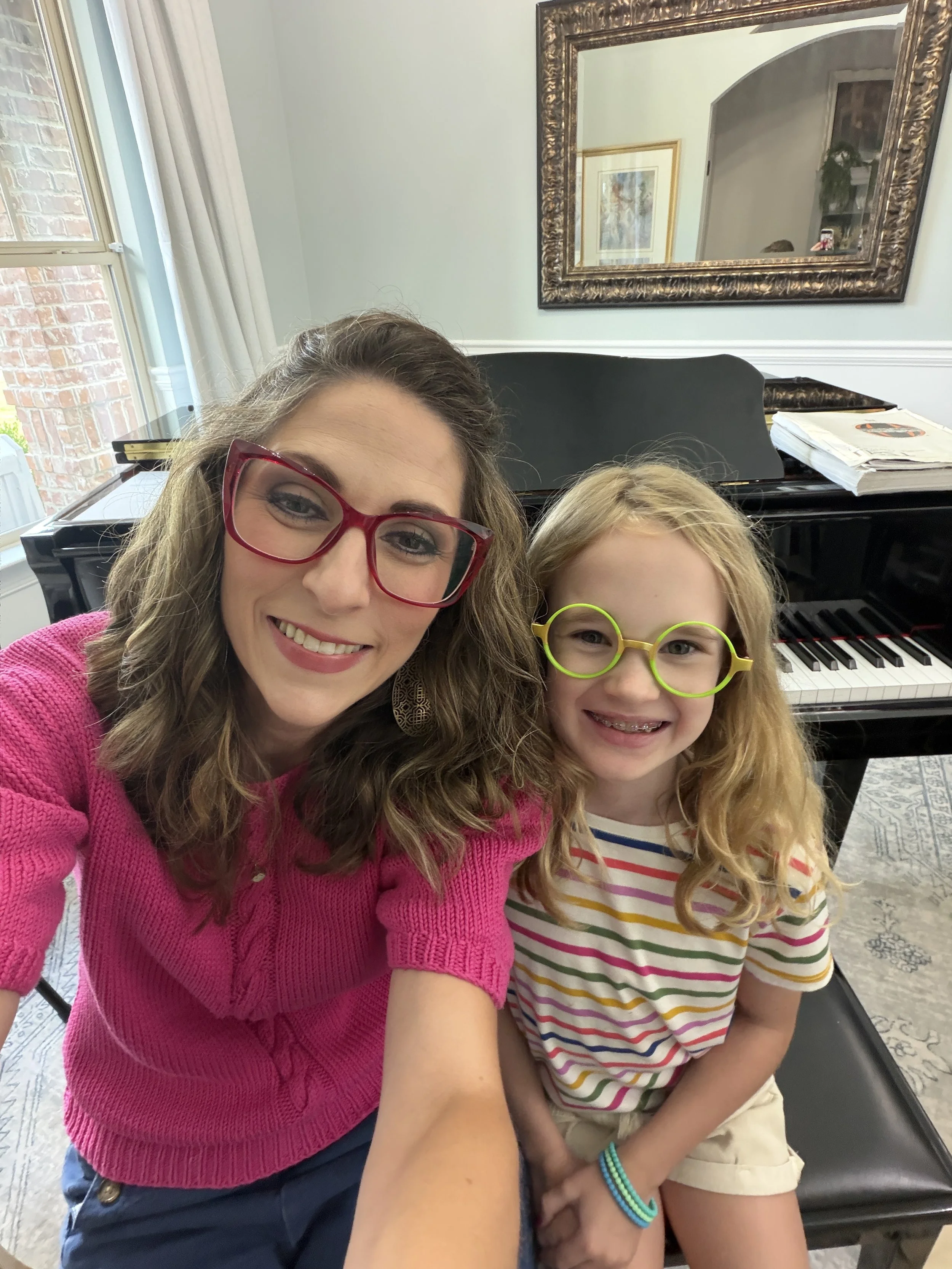 A woman with curly brown hair wearing red glasses and a pink sweater smiling for a selfie with a young girl with long hair, glasses, and a colorful striped shirt, sitting on a black piano bench in a room with a mirror on the wall, a grand piano, and sunlight coming through a window.