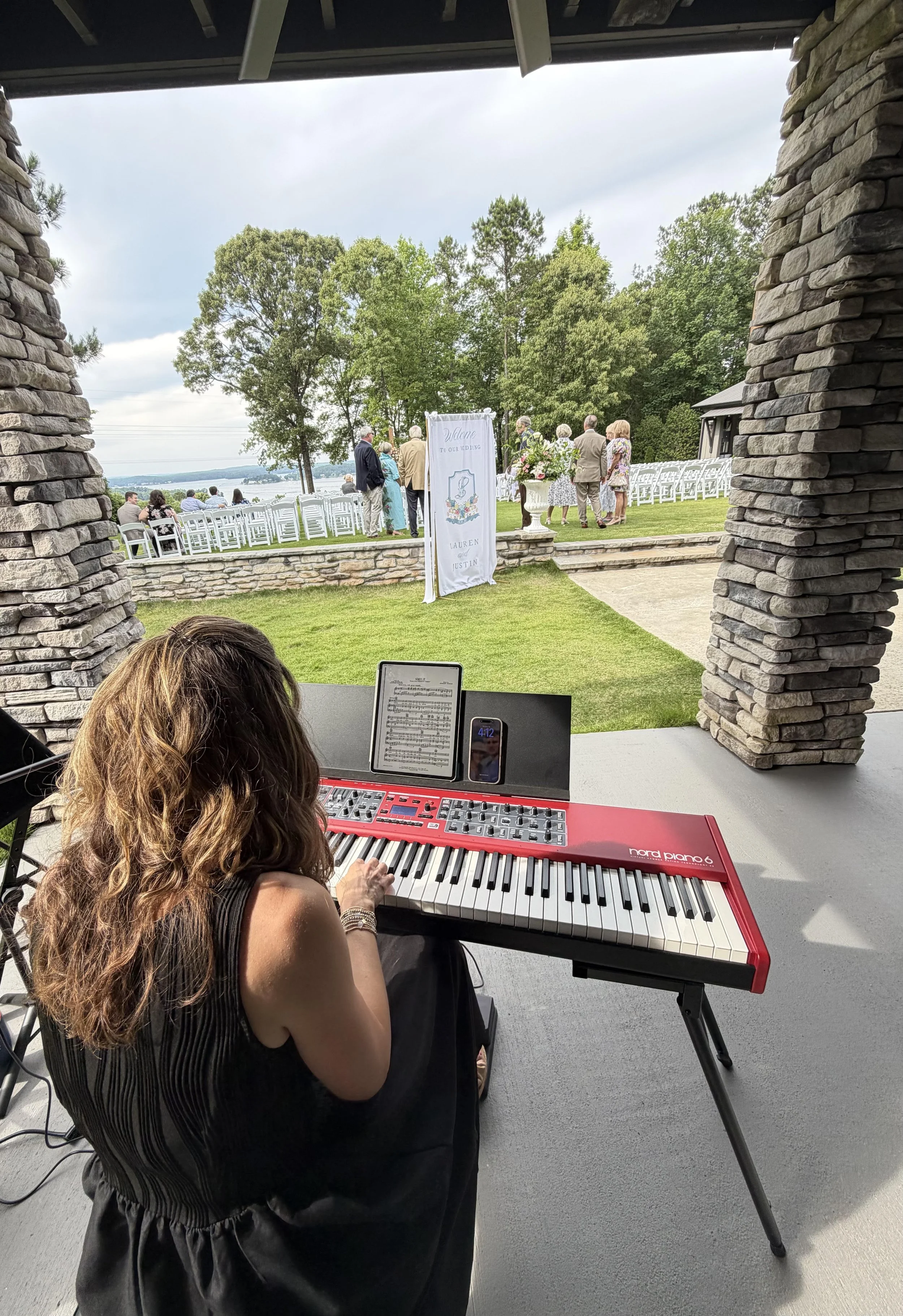 A woman playing a red Nord Piano 6 keyboard, with a music sheet on a stand, outdoors with a view of guests gathering near a lake, a sign welcoming guests, and large trees in the background during a wedding celebration.