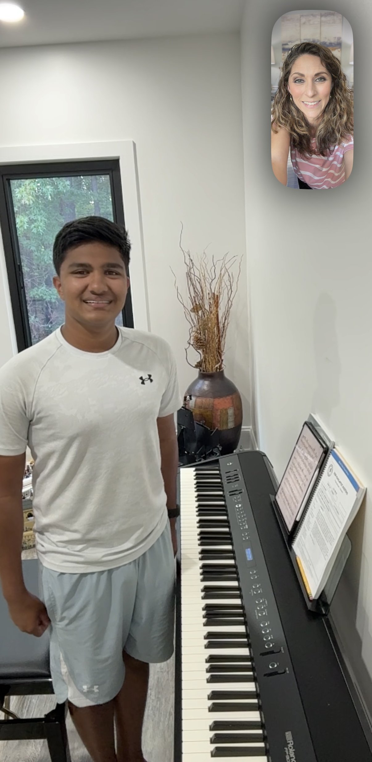 A young man standing beside a digital piano with sheet music on a stand and a woman smiling on a wall-mounted mirror behind him, in a bright room with a window.