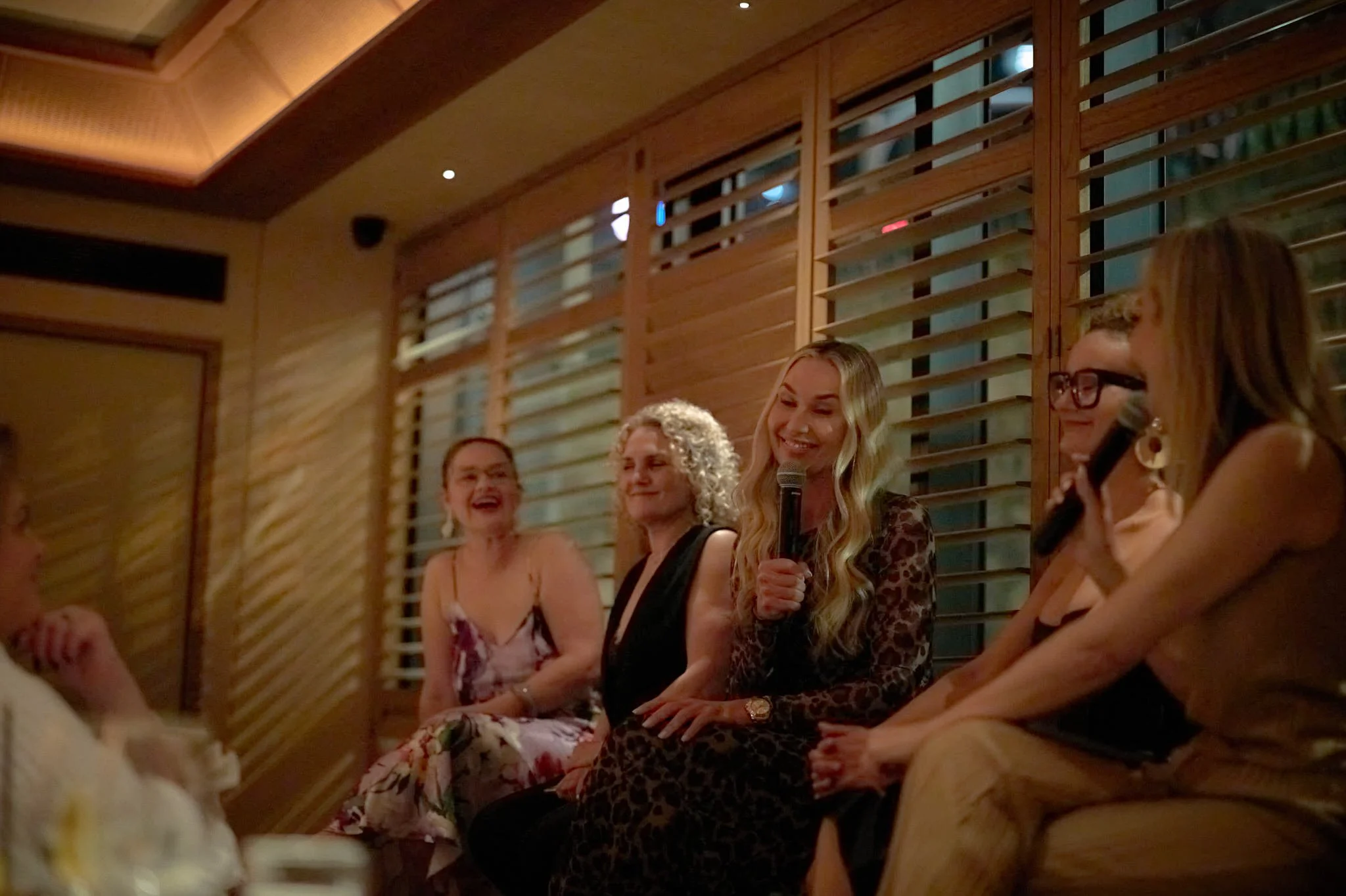 Group of women sitting indoors at a social gathering, smiling, some holding microphones, against a wooden wall with horizontal slats. Connecting lawyer mums  Leadership keynote speaker motivational speaker burnout recovery Rachael Karlyl