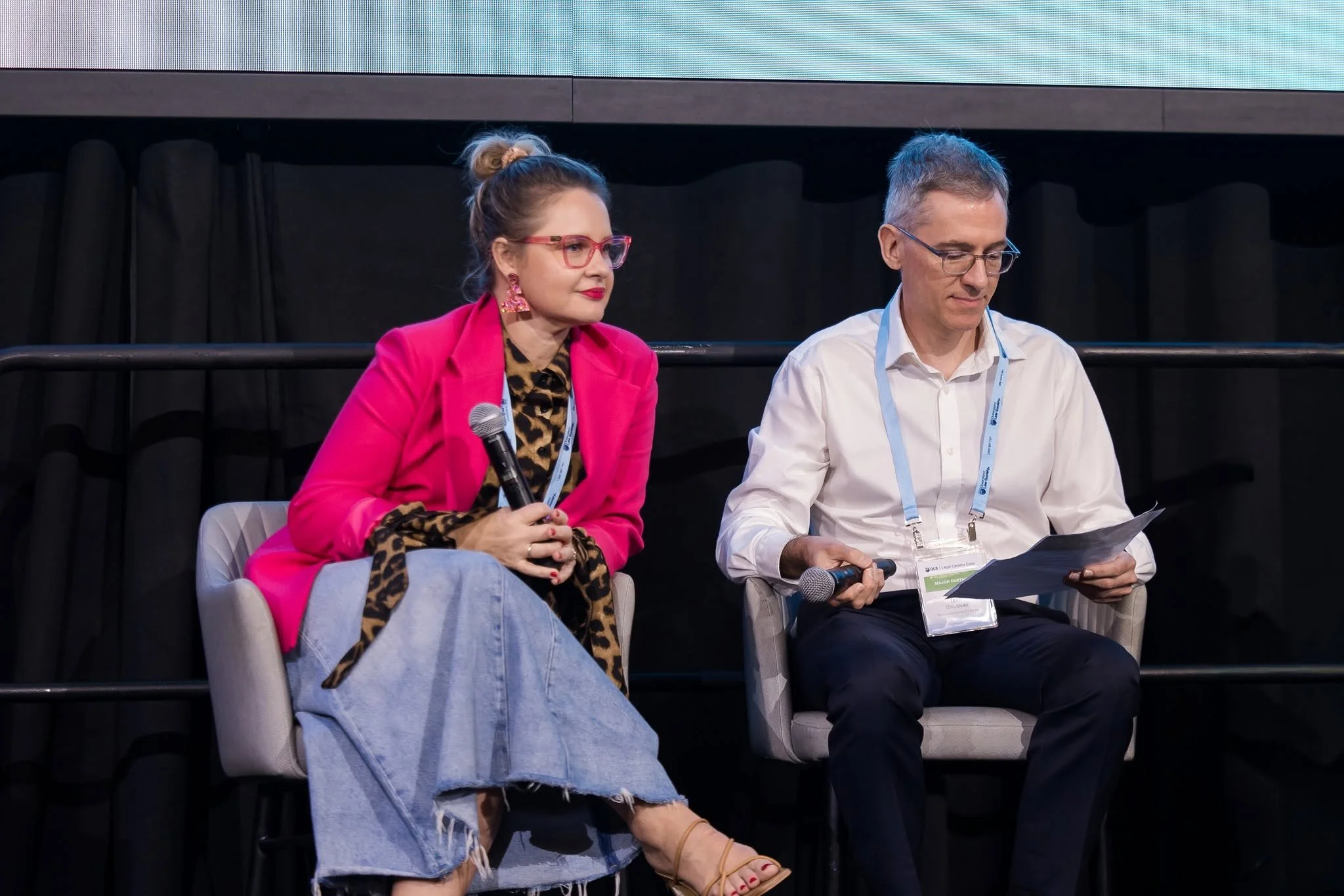 A woman with brown hair in a bun, wearing pink glasses, a bright pink blazer, leopard print shirt, and ripped jeans, sits holding a microphone. Next to her, a man with gray hair and glasses, in a white shirt with a conference badge, reads a paper.