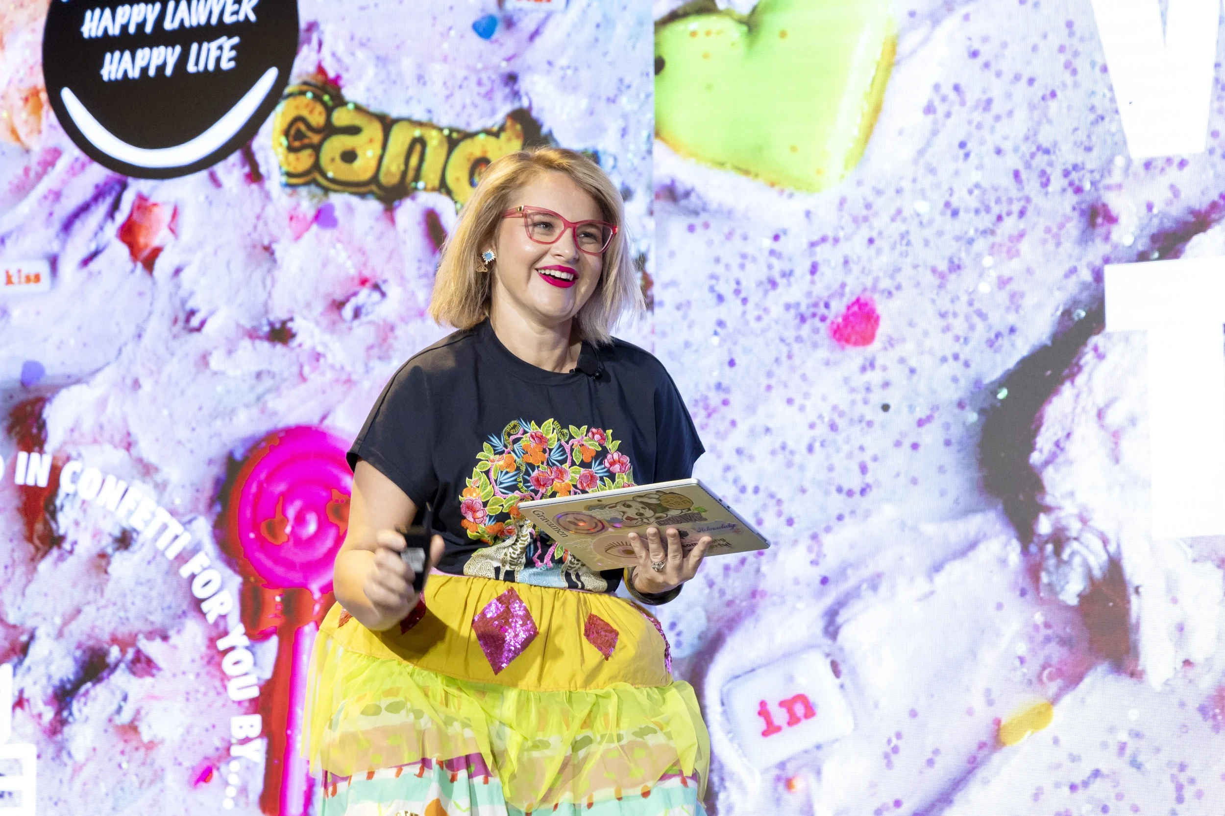 A woman with blonde hair, red glasses, and bright lipstick smiling while holding a tablet and a microphone, standing in front of a colorful, patterned backdrop with various phrases and images, including a large black circle with the words 'HAPPY LAWY