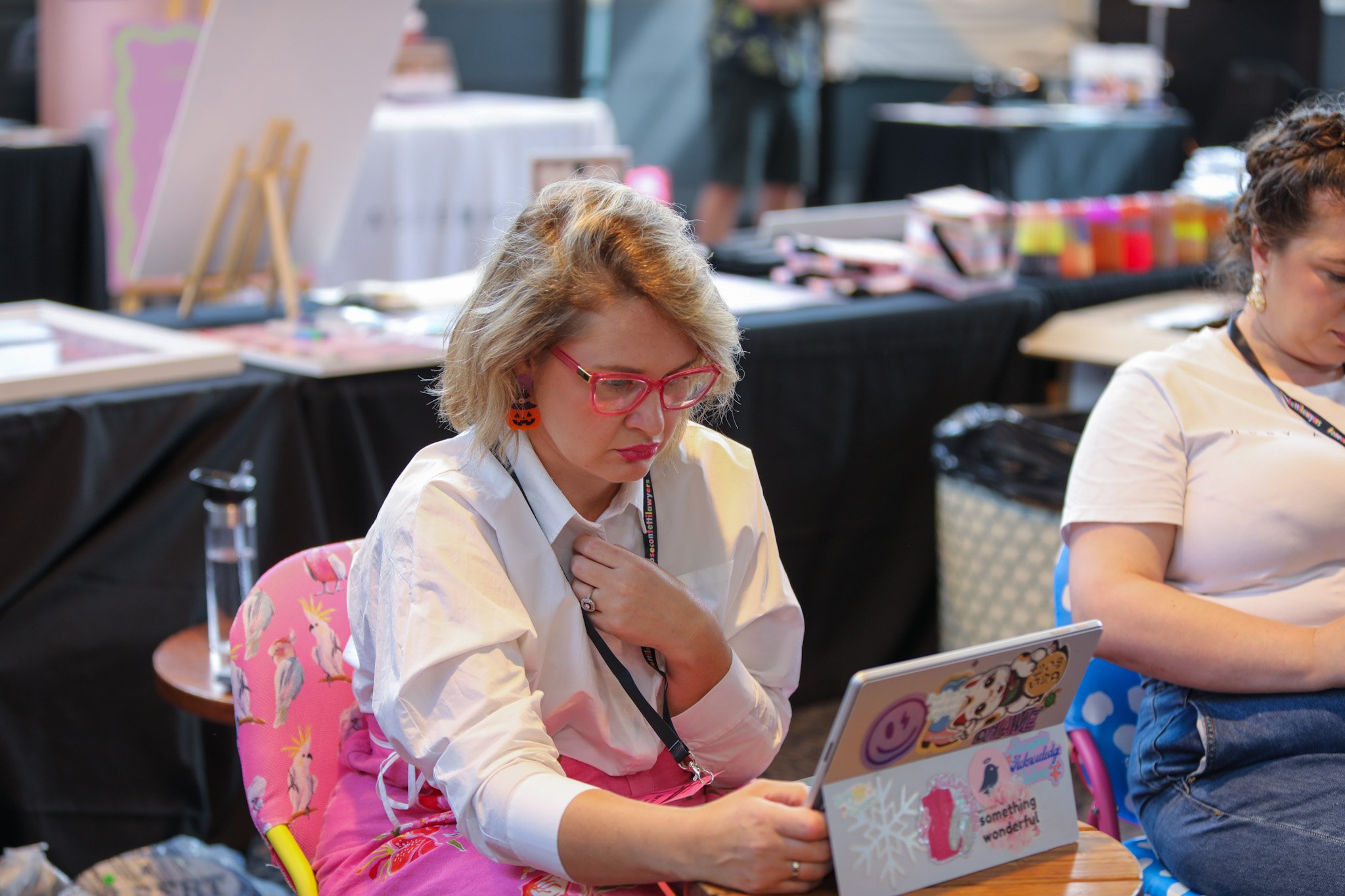 A woman with blonde hair, pink glasses, and orange pumpkin earrings is sitting at a table, looking at a tablet. She is wearing a white shirt and pink pants with a unicorn pattern. Other people are sitting nearby, and there are tables with art supplie