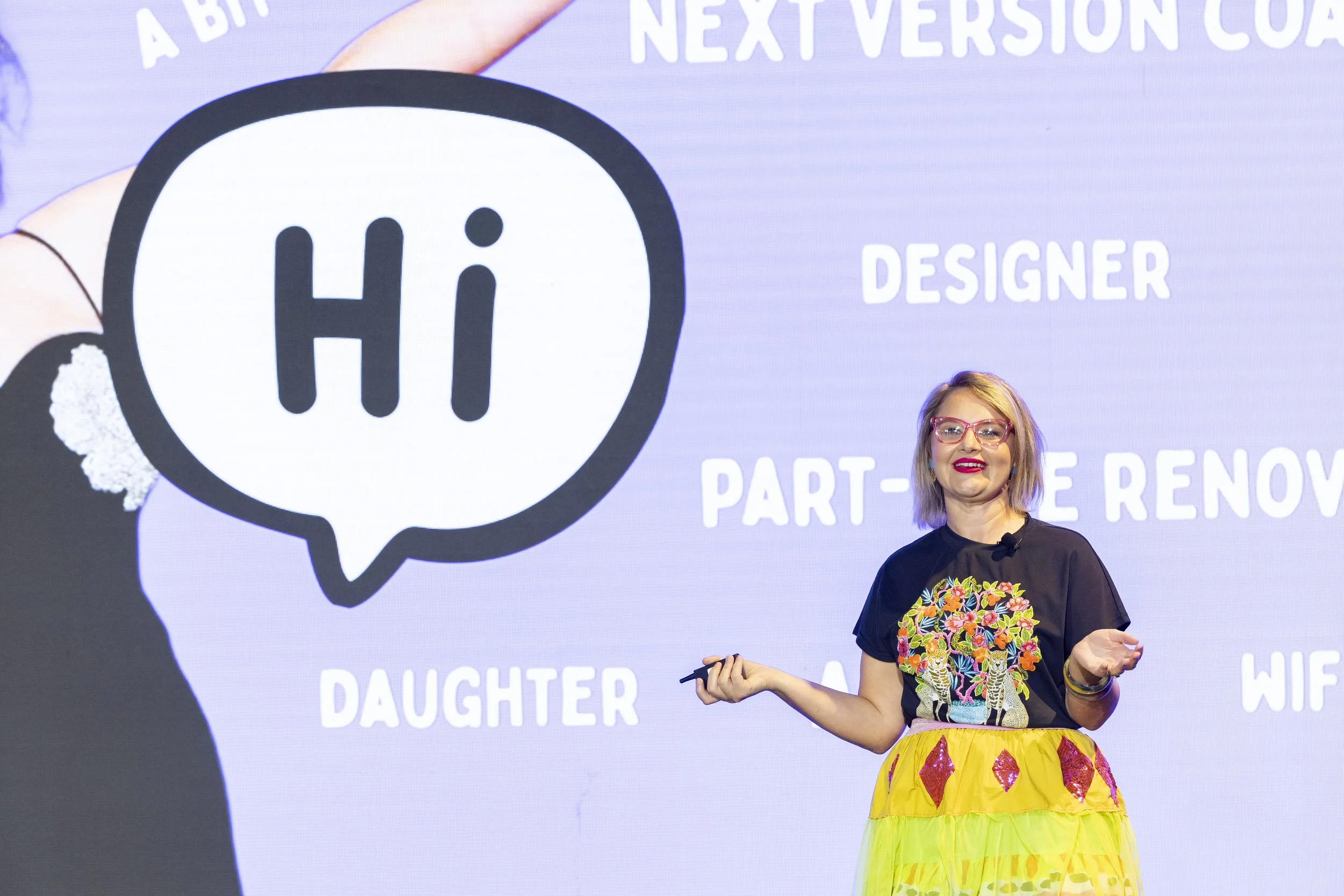 A woman with glasses and colorful attire, wearing a yellow skirt with pink diamonds on it, standing in front of a presentation screen with a large logo and text.  Leadership keynote speaker motivational speaker burnout recovery Rachael Karlyl