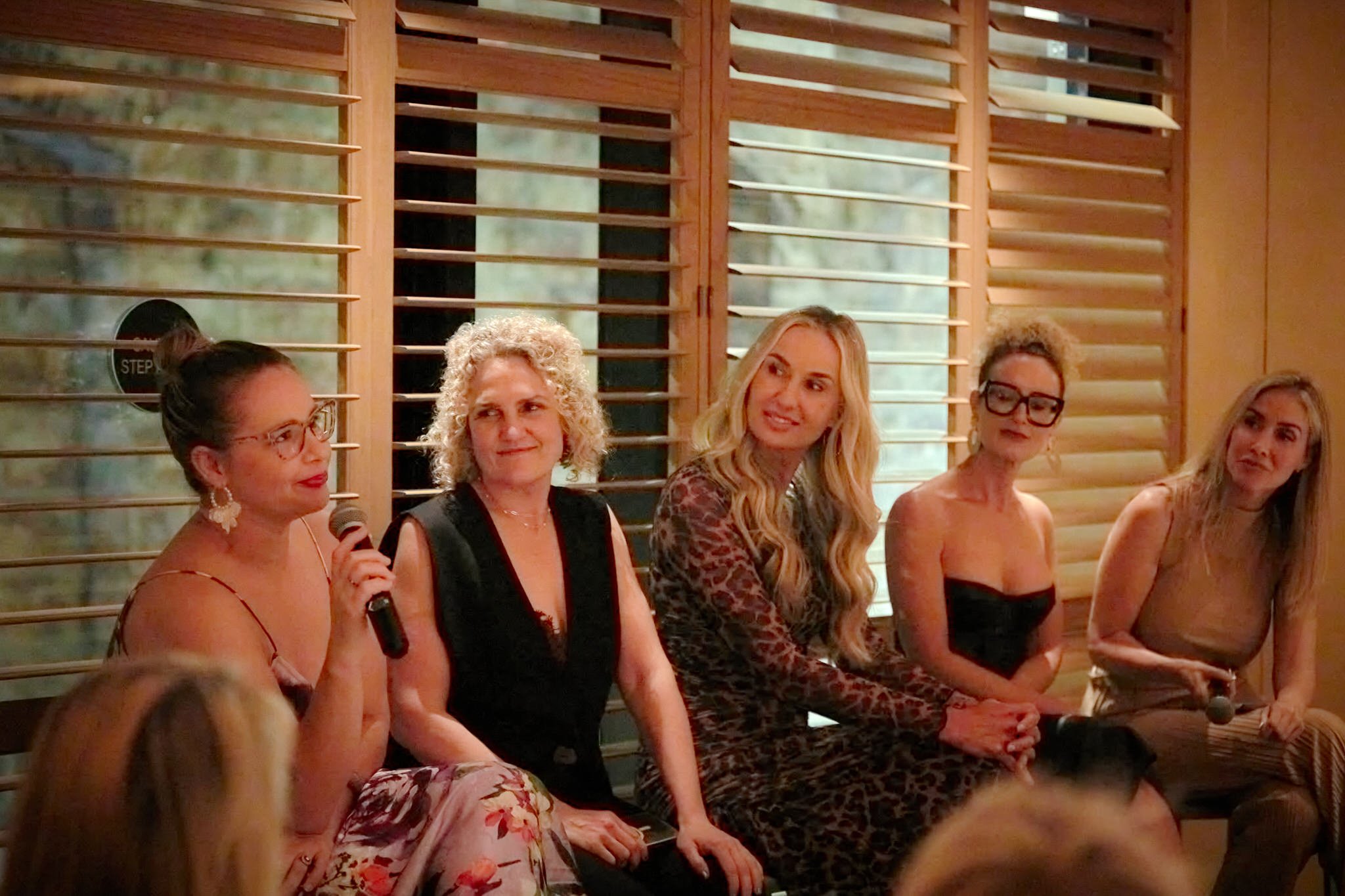 Group of women sitting at a panel or discussion inside a room with wooden window shutters, appears to be a formal or semi-formal event.  Leadership keynote speaker motivational speaker burnout recovery Rachael Karlyl connecting lawyer mums