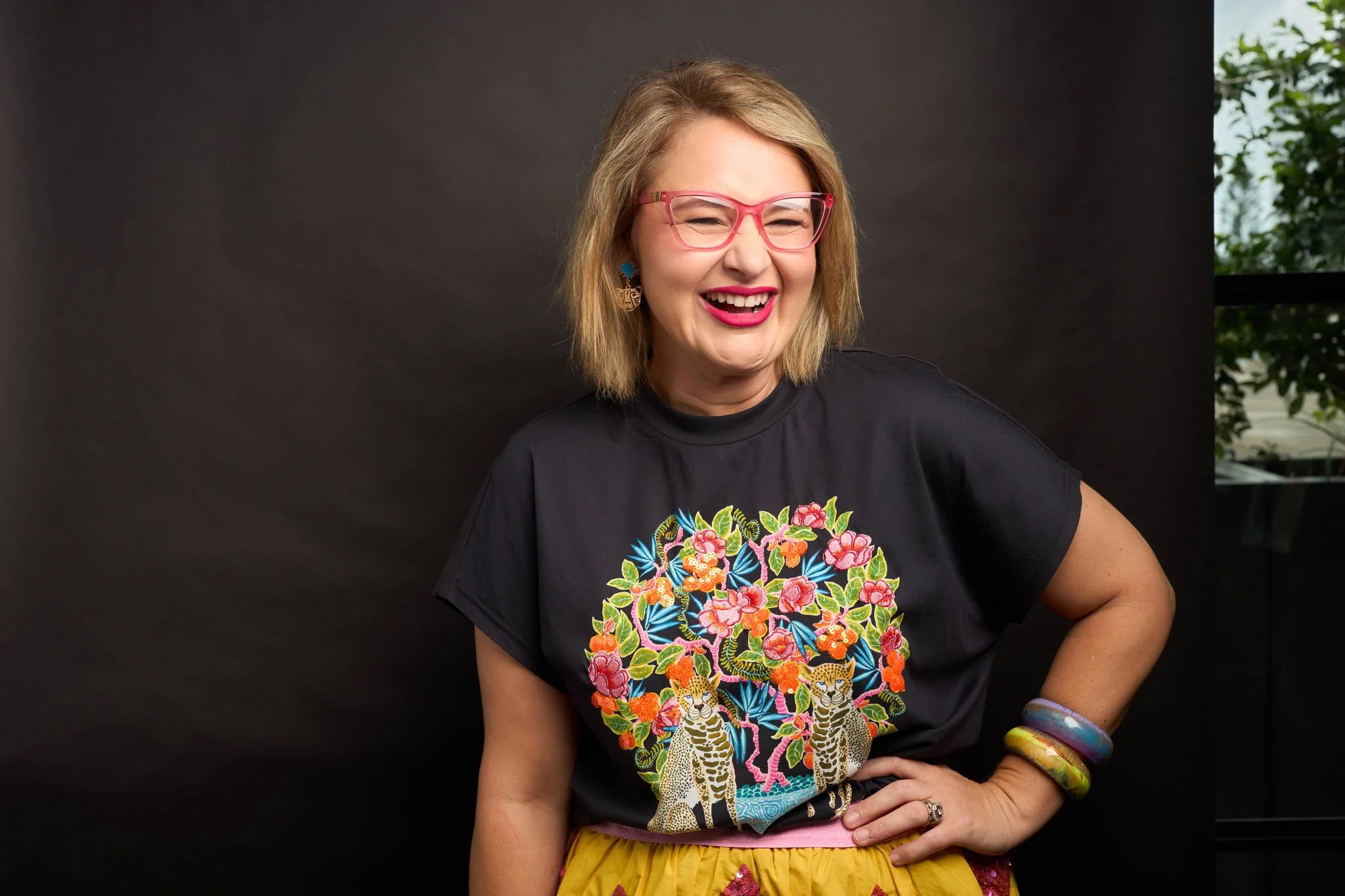 A smiling woman with blonde hair wearing pink glasses, a black floral shirt, and yellow skirt stands against a dark background, with greenery visible on the right side.