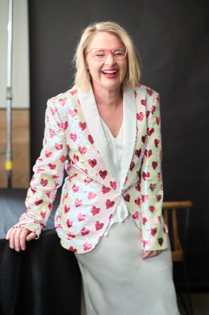 Woman with blonde hair, pink glasses, and a heart-patterned blazer, smiling and laughing indoors.  Leadership keynote speaker motivational speaker burnout recovery Rachael Karlyl