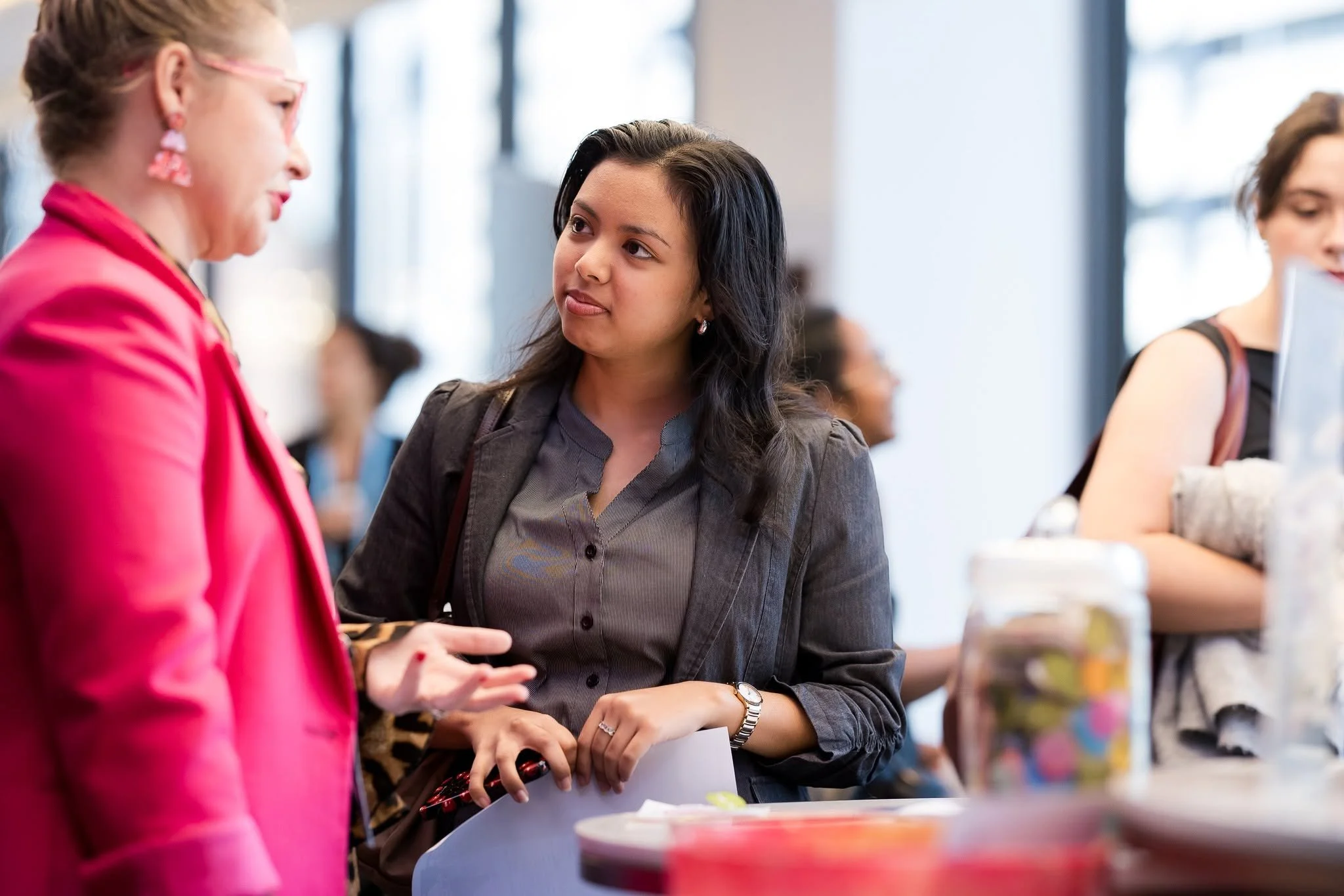 Two women engaged in conversation behind a table at an indoor event with large windows. One woman has blond hair, wearing pink, and the other has dark hair, wearing a gray jacket.