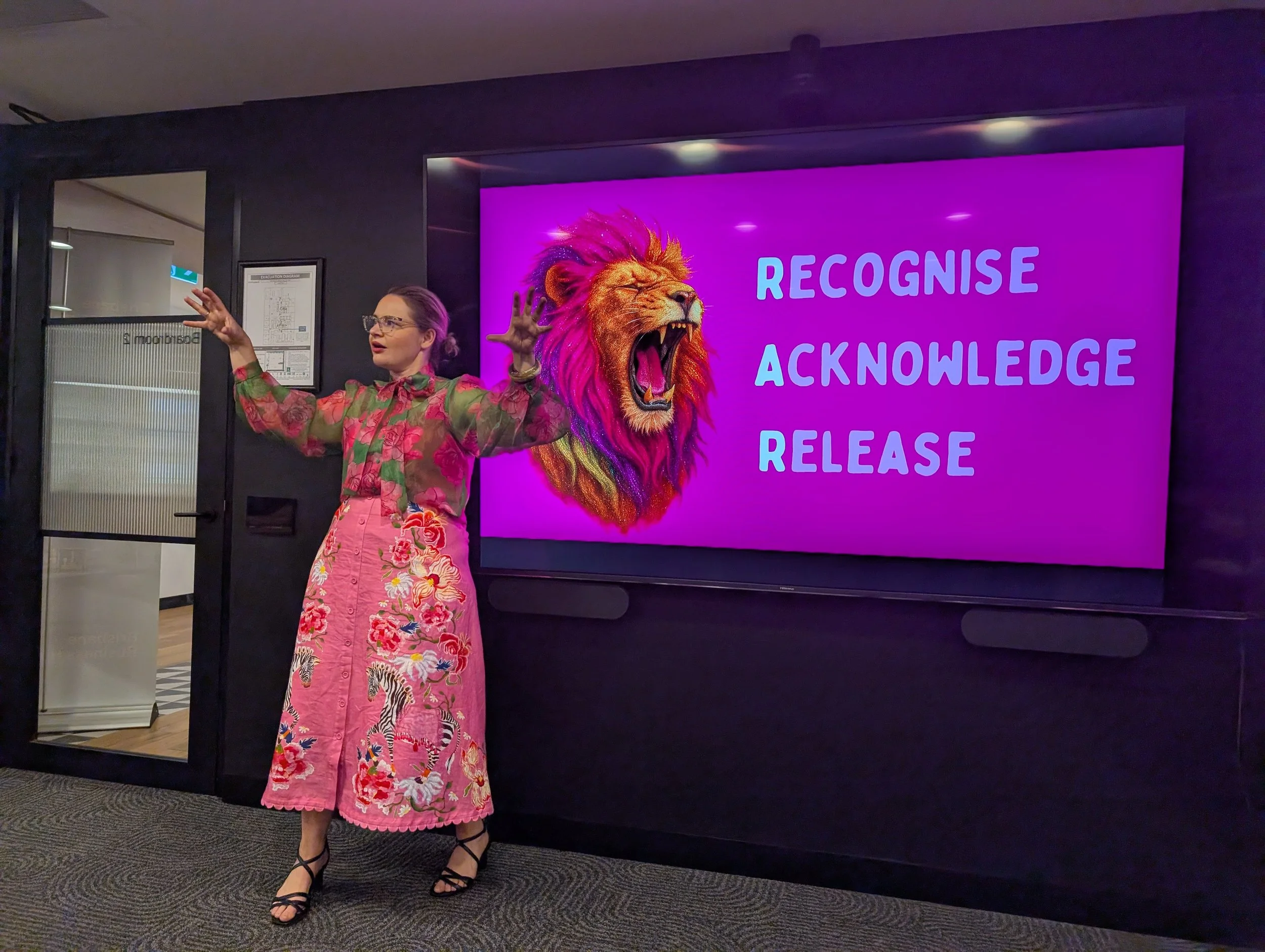 A woman in a pink floral skirt and floral blouse stands in front of a digital sign displaying a lion with a colorful mane and the words 'Recognise, Acknowledge, Release' in purple. She is gesturing with her hands and speaking.  Leadership burnout