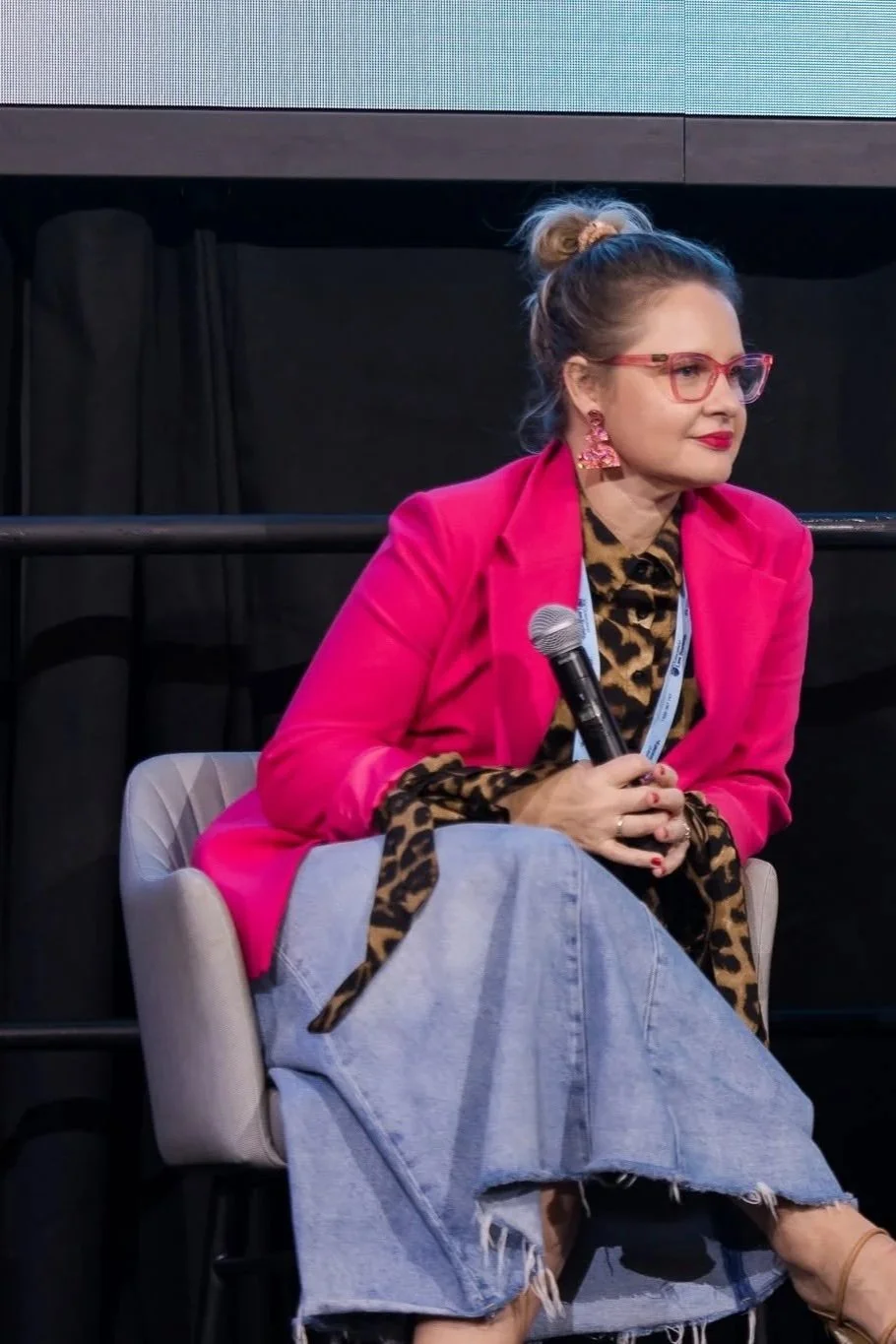Woman in pink blazer with leopard print scarf and jeans sitting with a microphone in her hands at an event. Leadership keynote speaker motivational speaker Rachael Karlyl dress for the job you want enclothed cognition burnout recovery