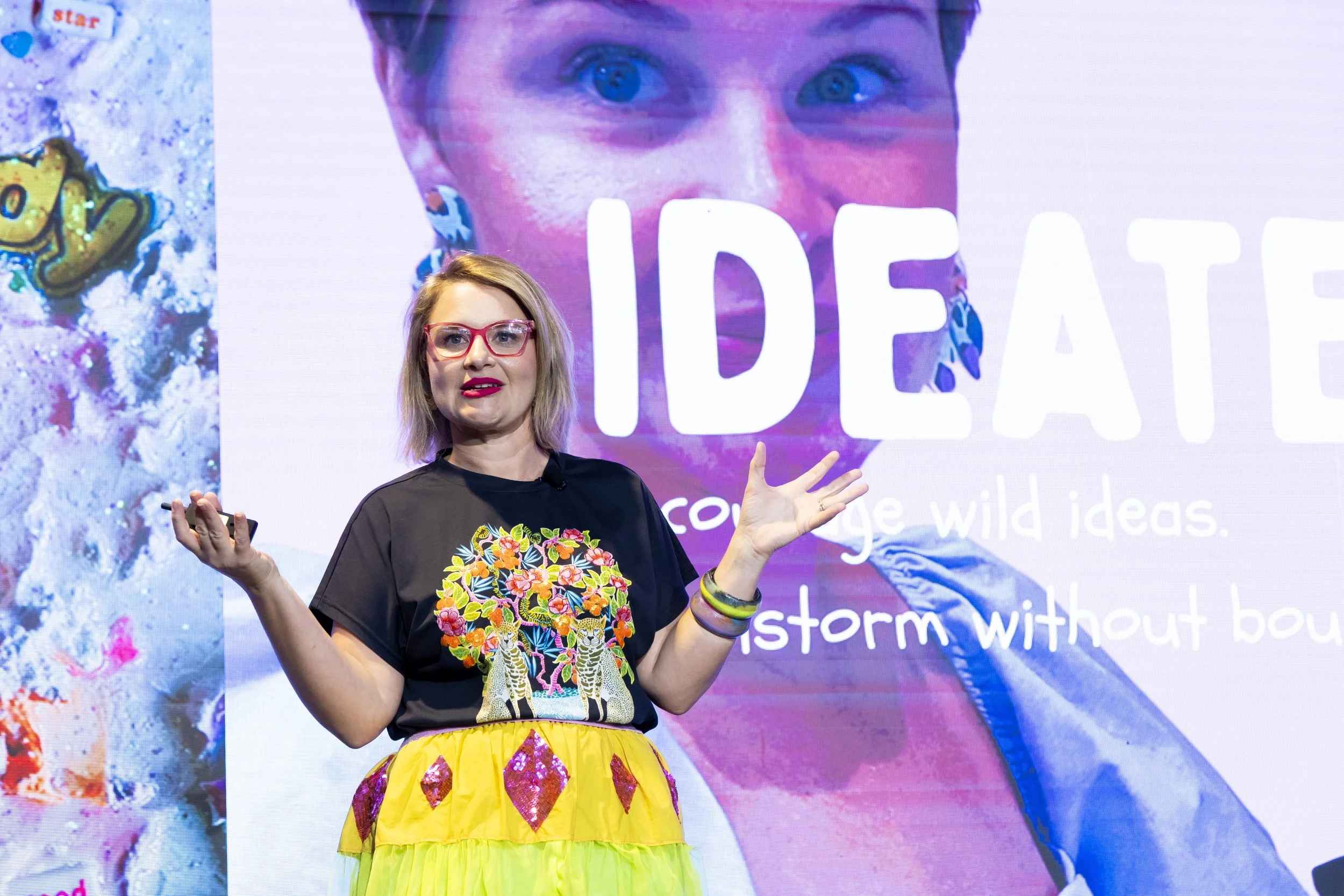 A woman with blonde hair, red glasses, and colorful earrings giving a presentation on stage. She is wearing a black shirt with a vibrant embroidered tiger design and a multi-colored skirt. Behind her is a large screen displaying a close-up of her fac