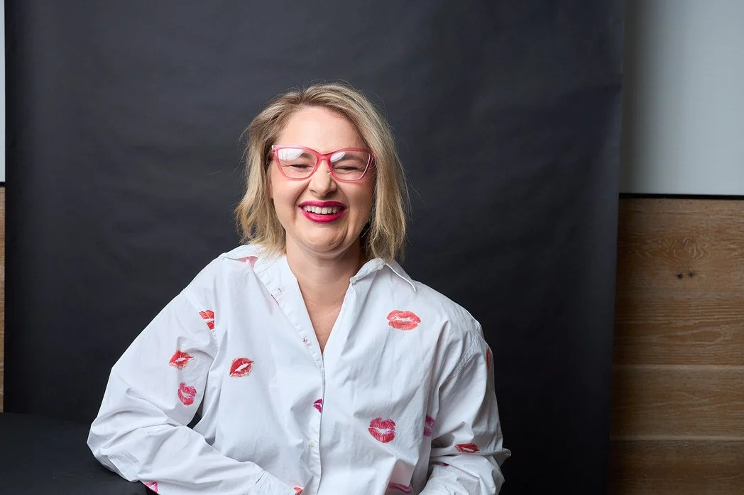 Woman with blonde hair and pink glasses smiling, wearing a white shirt with pink lipstick kiss prints, against a dark and wooden background.  Leadership keynote speaker motivational speaker burnout recovery Rachael Karlyl