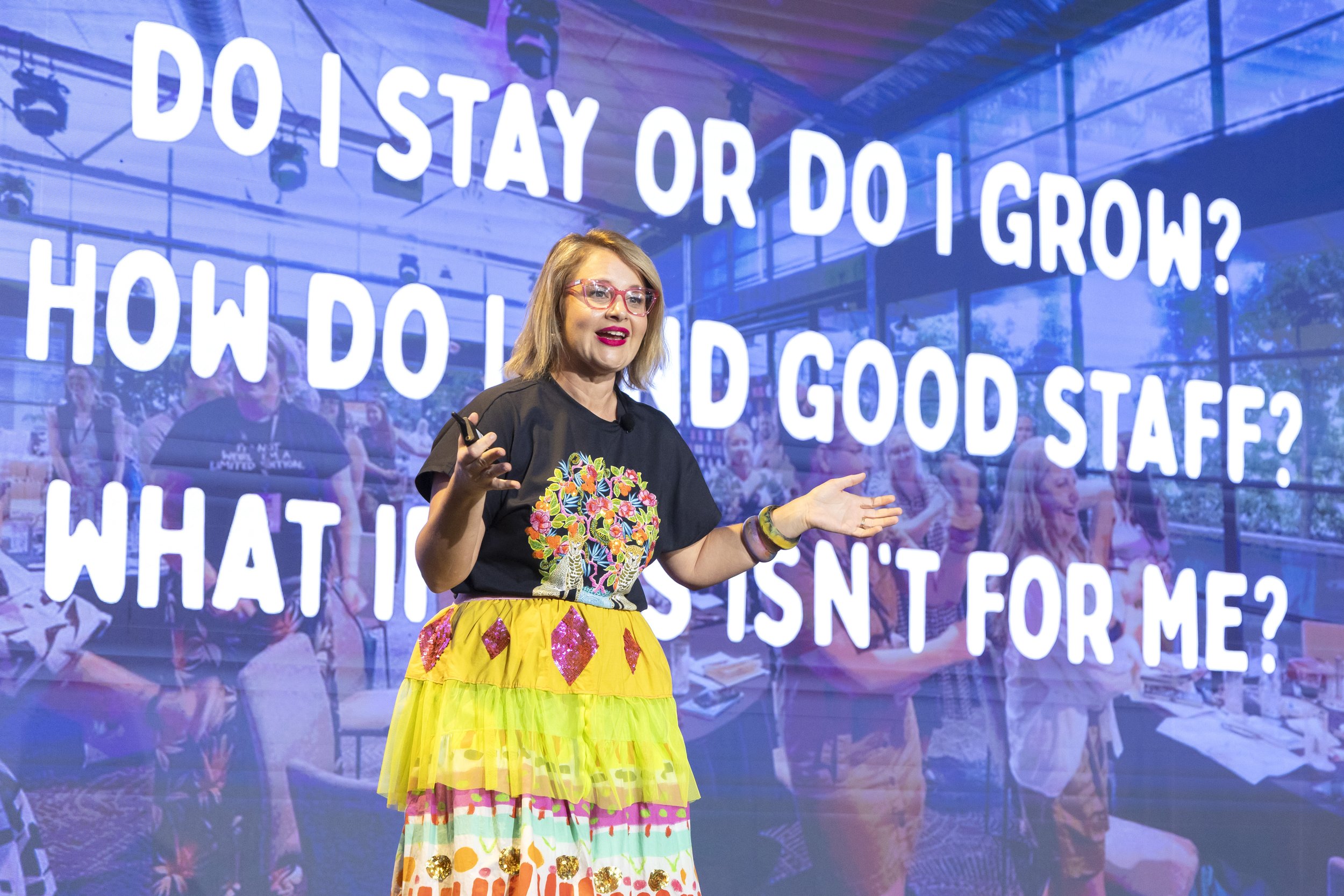 A woman with blonde hair and colorful glasses giving a presentation on a stage with a large screen behind her. The screen displays the question 'Do stay or do I grow? How do I do?' in large white letters. The woman is wearing a black top with a color